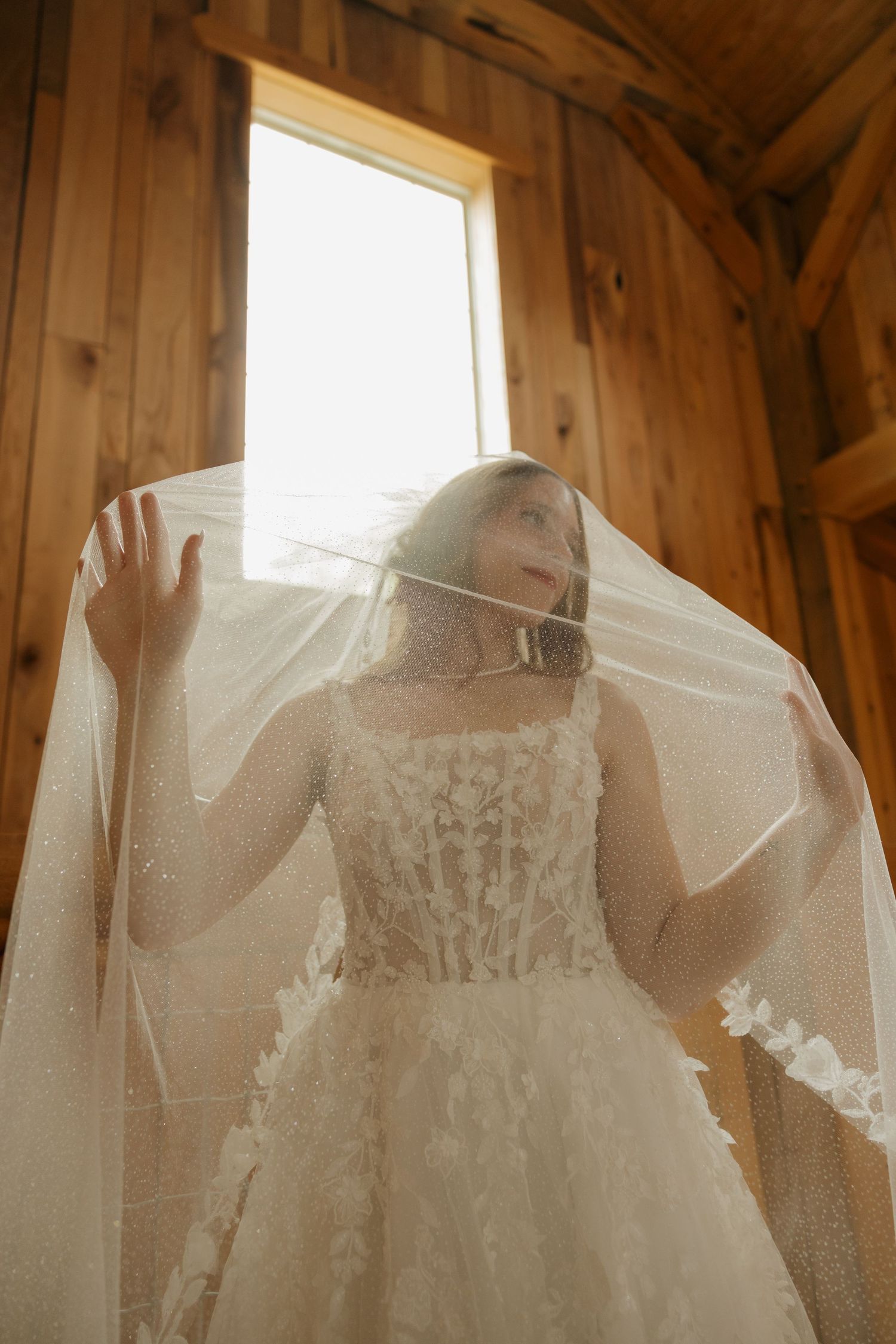 Bride in lace wedding gown and veil poses by a window in a warm wooden interior.