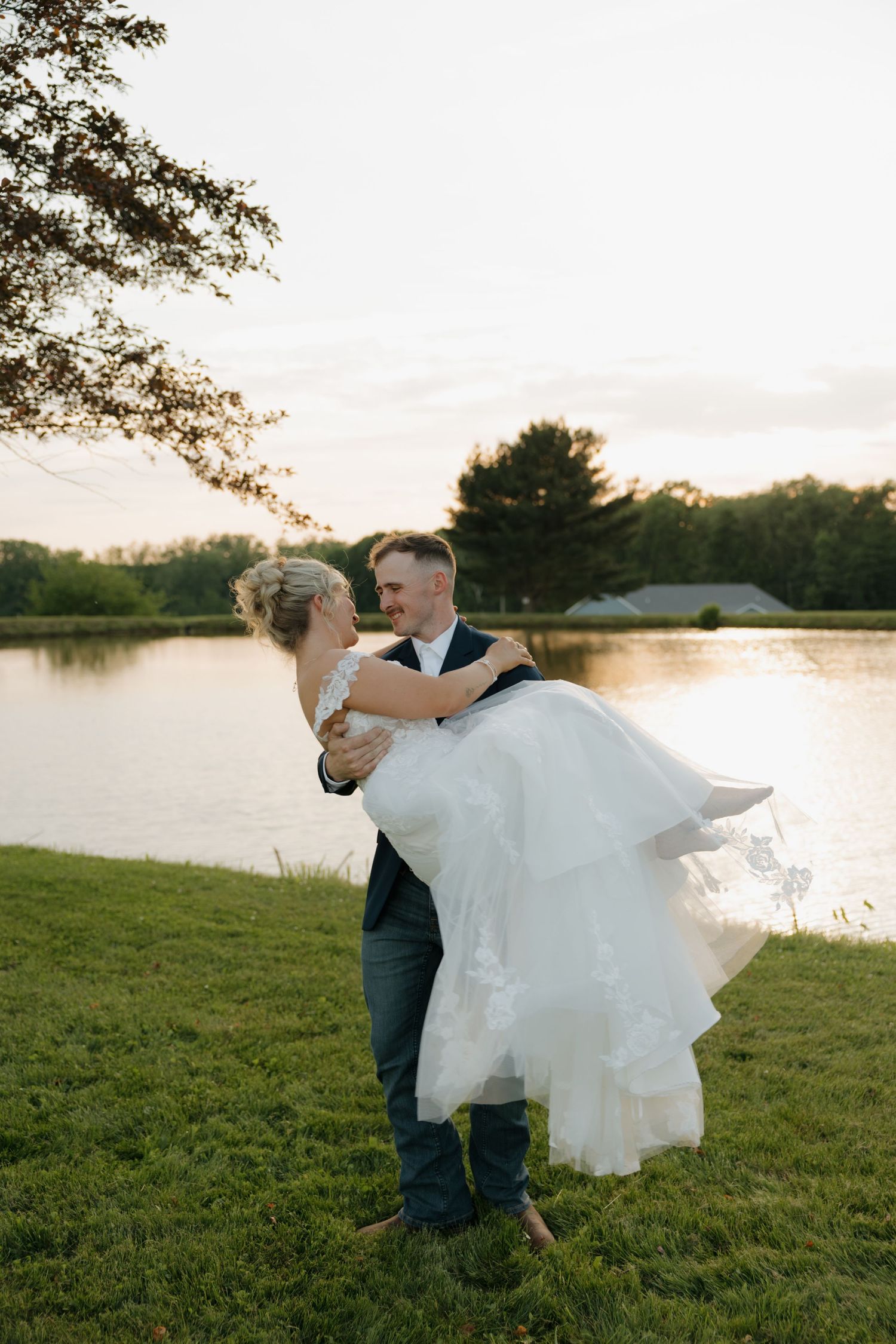 Groom lifting bride in white gown by a serene lake at sunset, sharing a joyful moment on lush green grass.