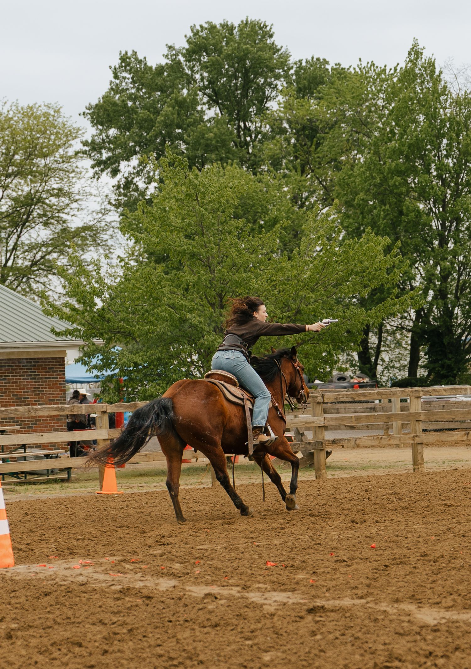 A woman rides a brown horse bareback in a dirt arena, arms outstretched, with green trees and wooden fencing in the background.