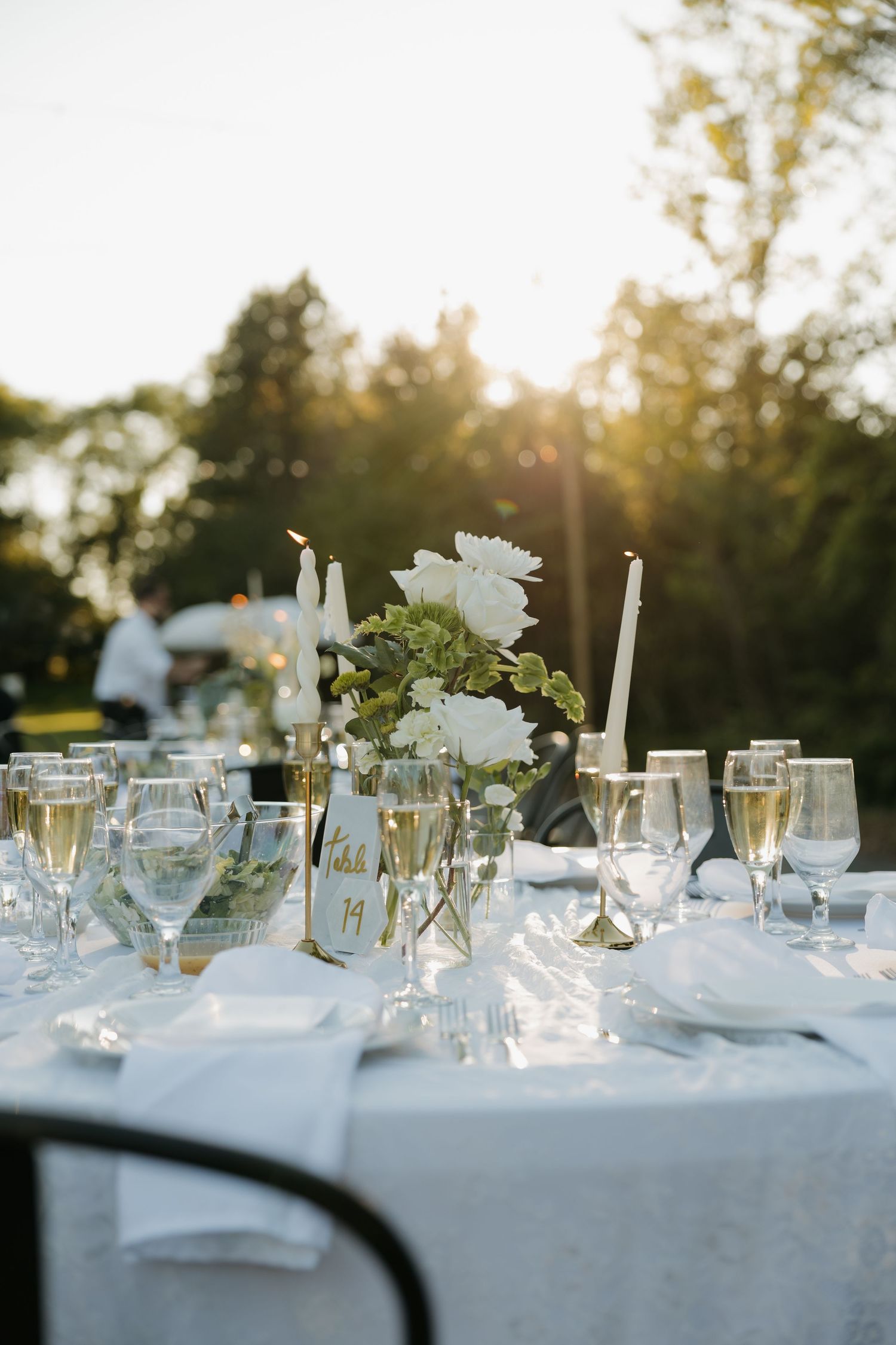 Elegant outdoor wedding reception table set with white florals, candles, champagne glasses, and a gold table number card.