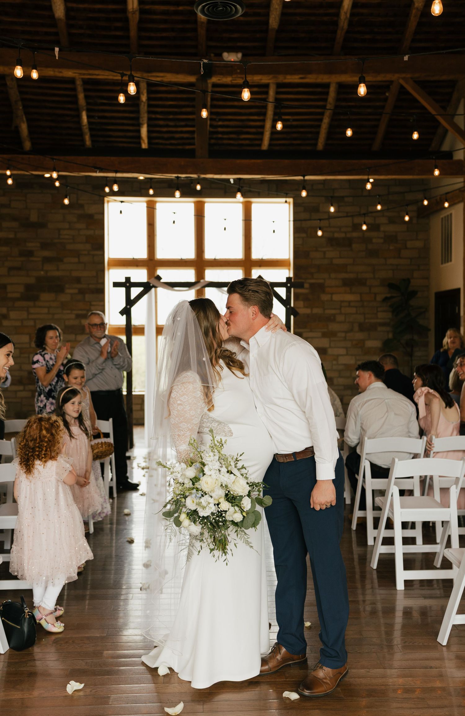 Bride and groom share first kiss at rustic indoor wedding venue with string lights and white chairs.