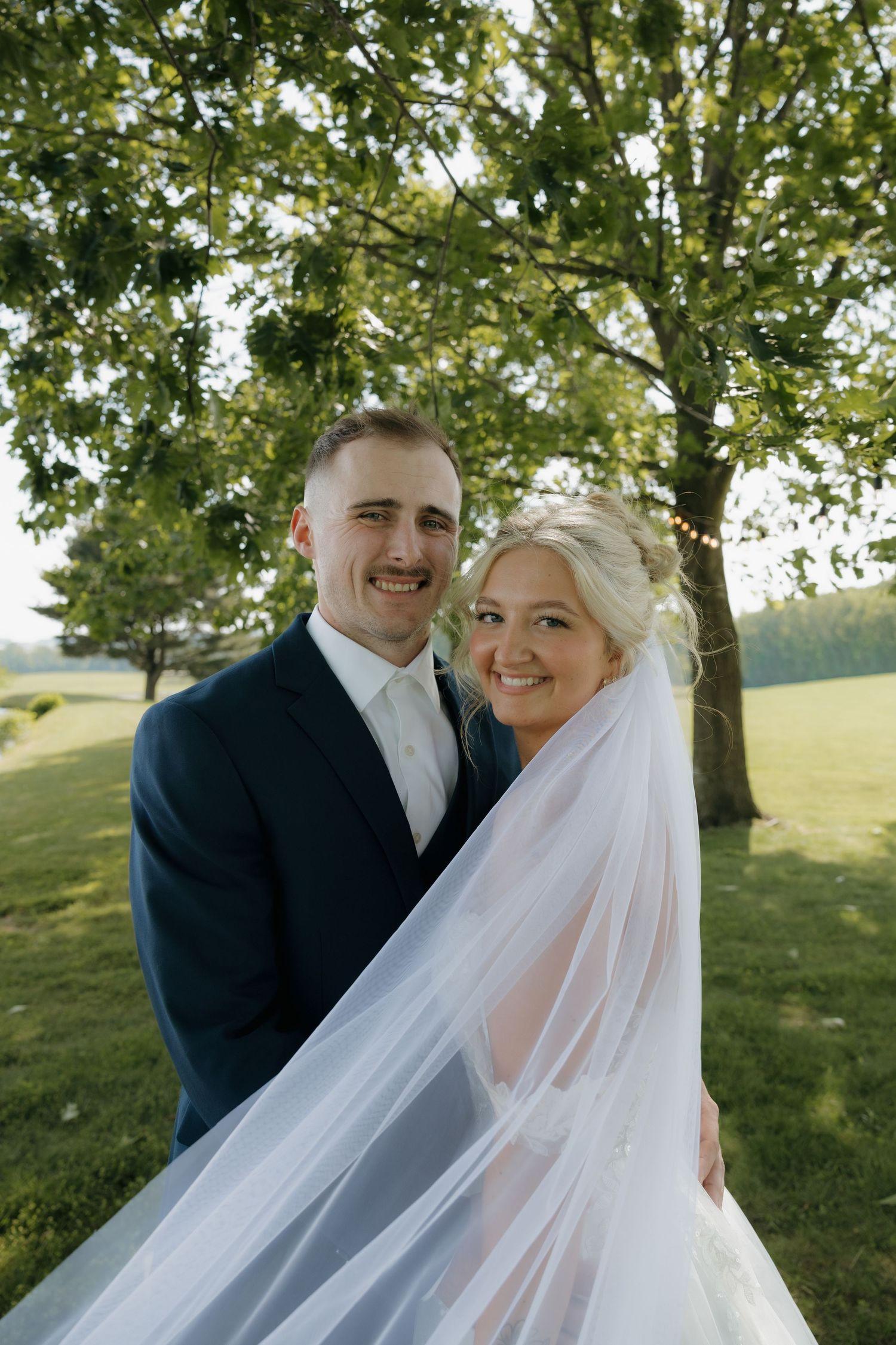 Bride and groom smiling outdoors under green tree, bride wearing white veil, groom in navy suit, golden hour lighting.