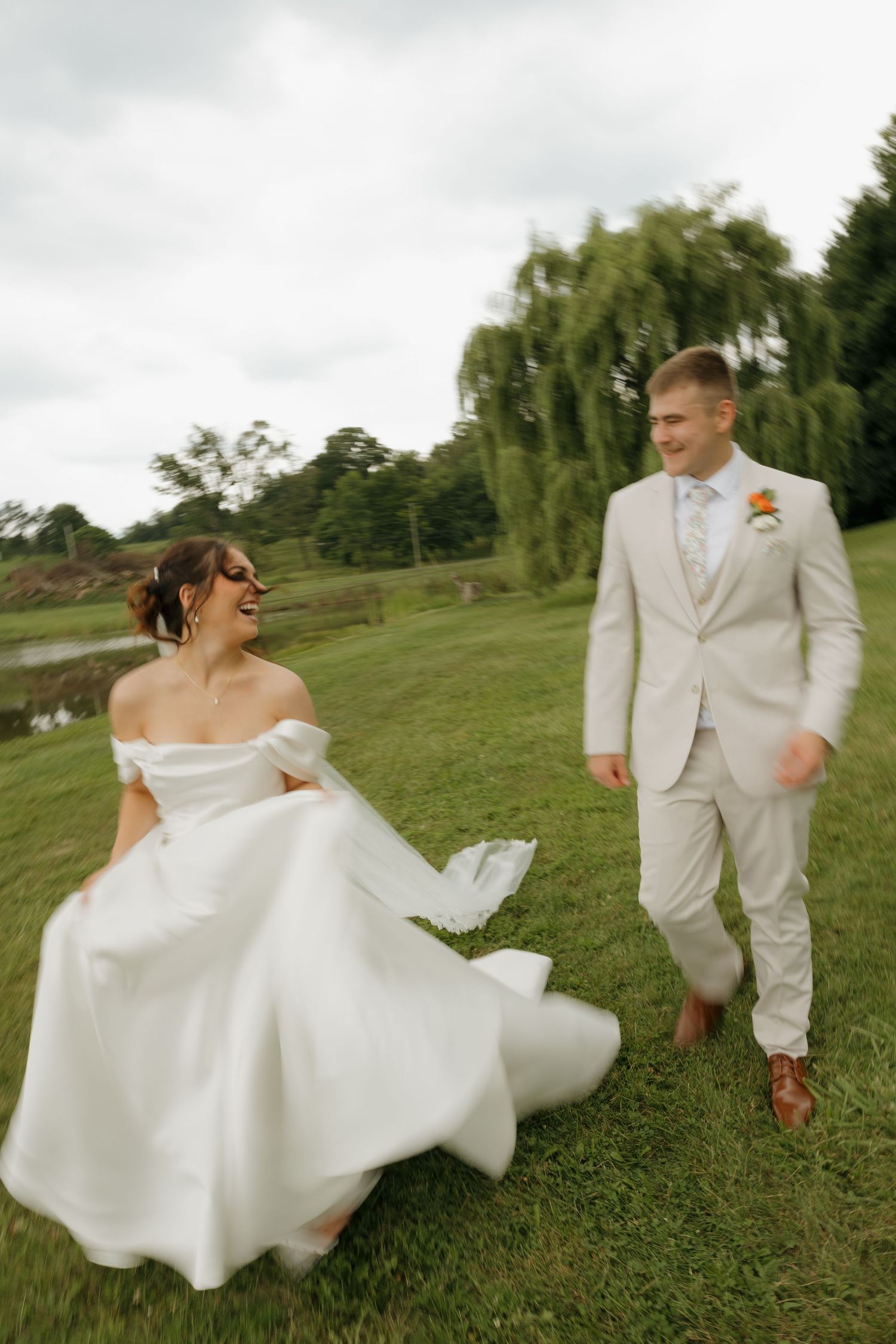Bride in white off-shoulder gown and groom in beige suit walk joyfully on green lawn with willow tree backdrop.
