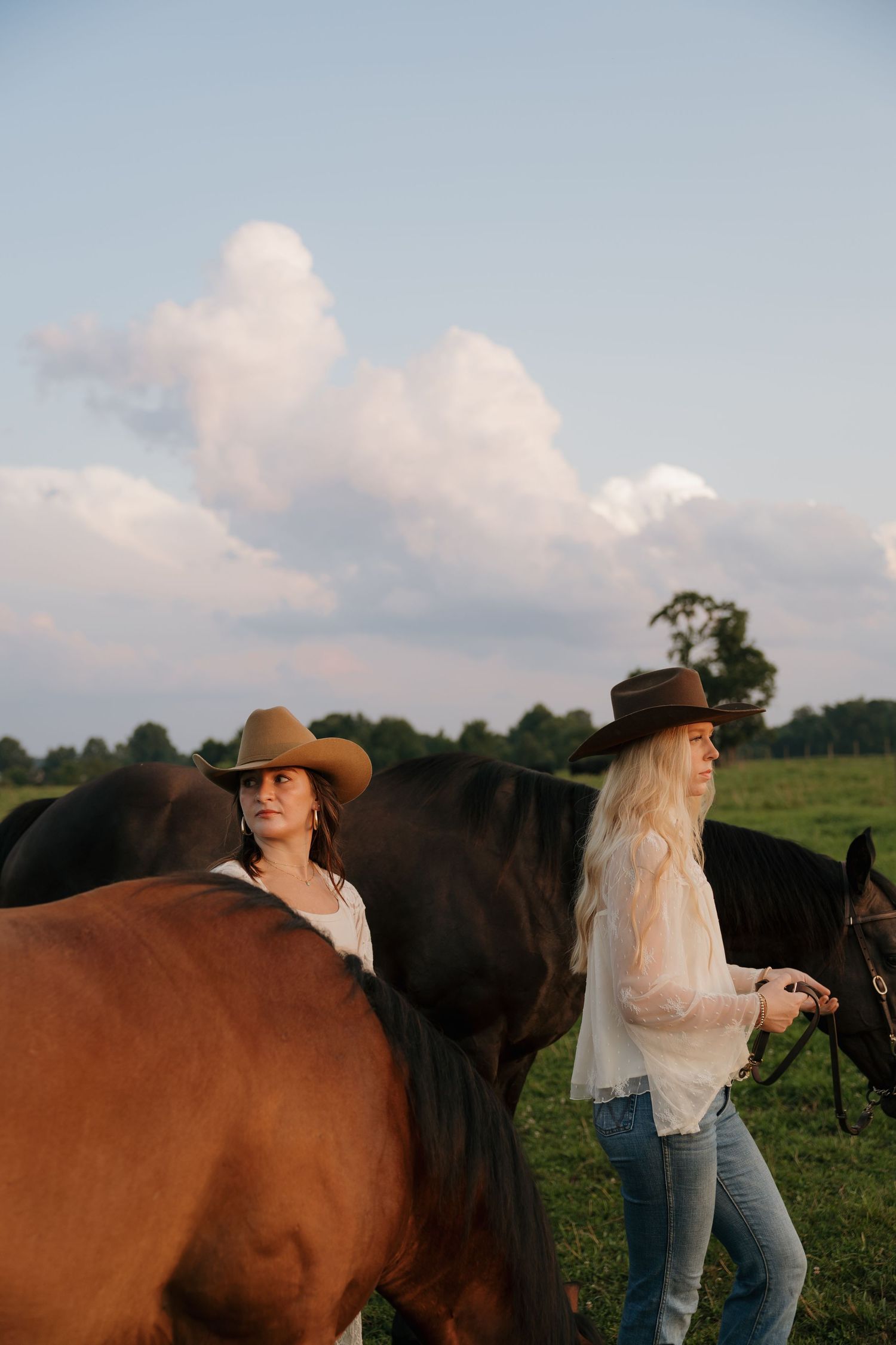 Two women in cowboy hats and white blouses walk alongside dark horses in a green field under a cloudy sky.