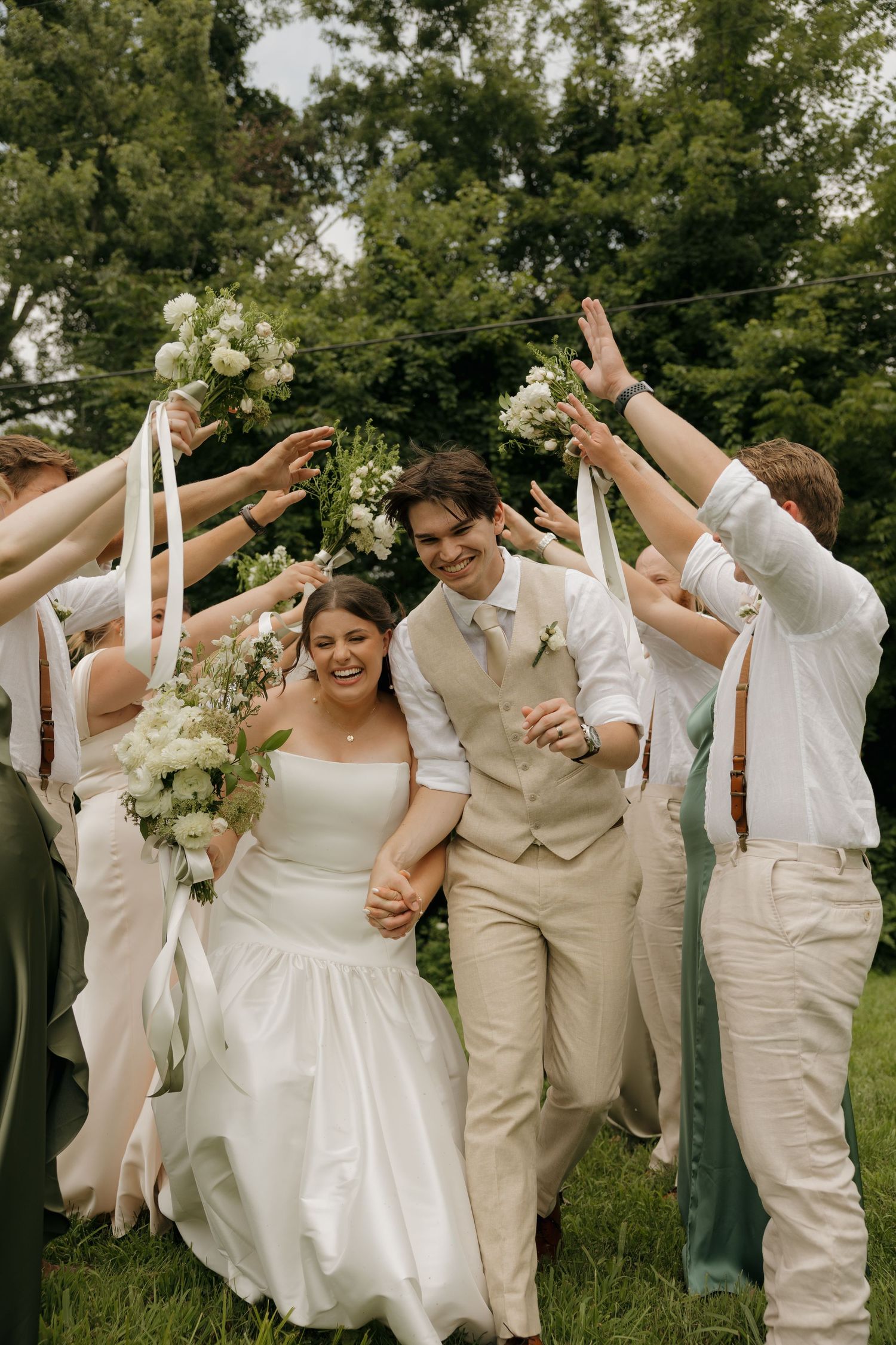 A joyful bride and groom walk through an arch of raised bouquets held by their wedding party outdoors.