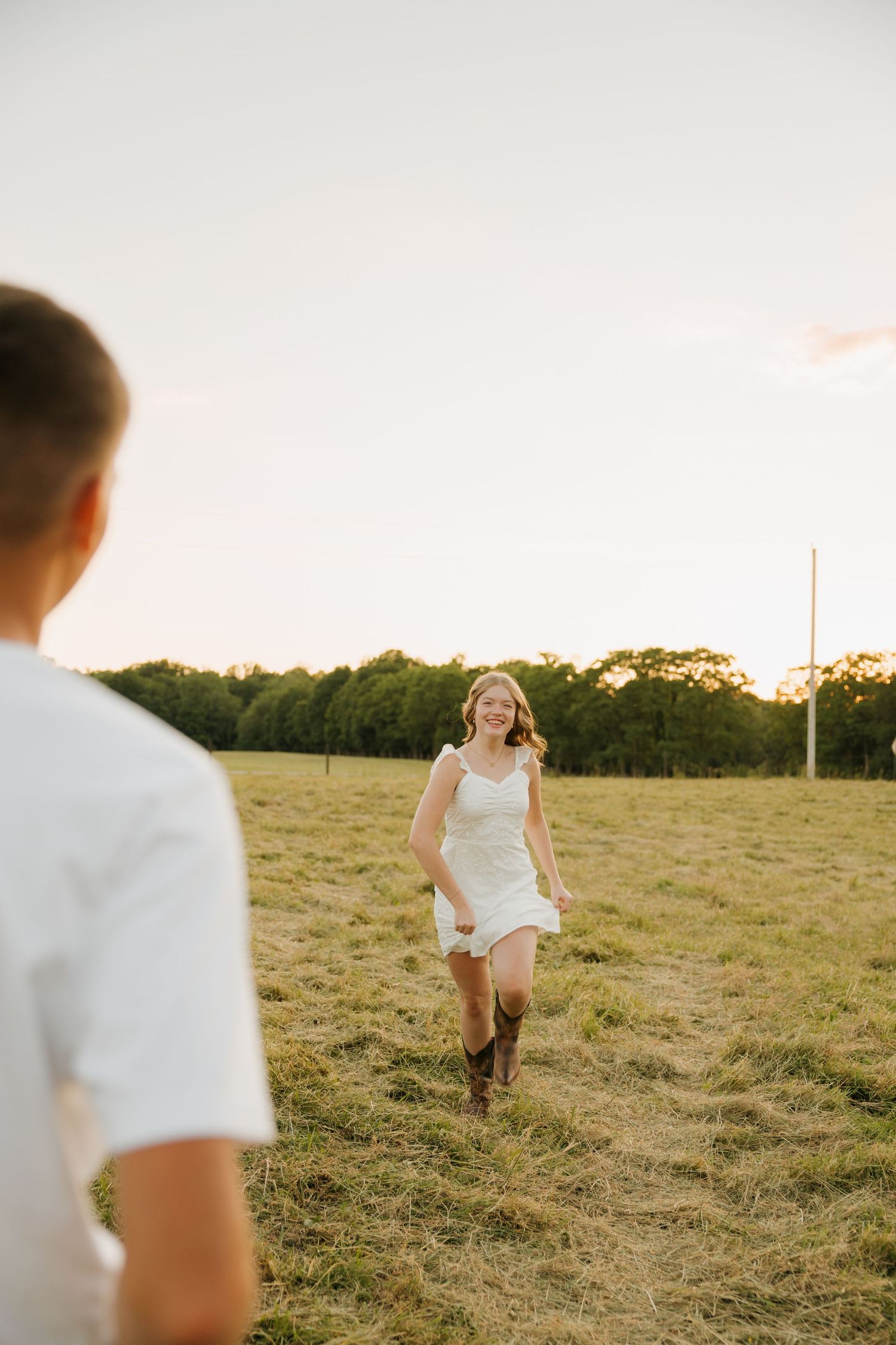 Young woman in white dress and cowboy boots running through a sunlit field toward her partner at golden hour.