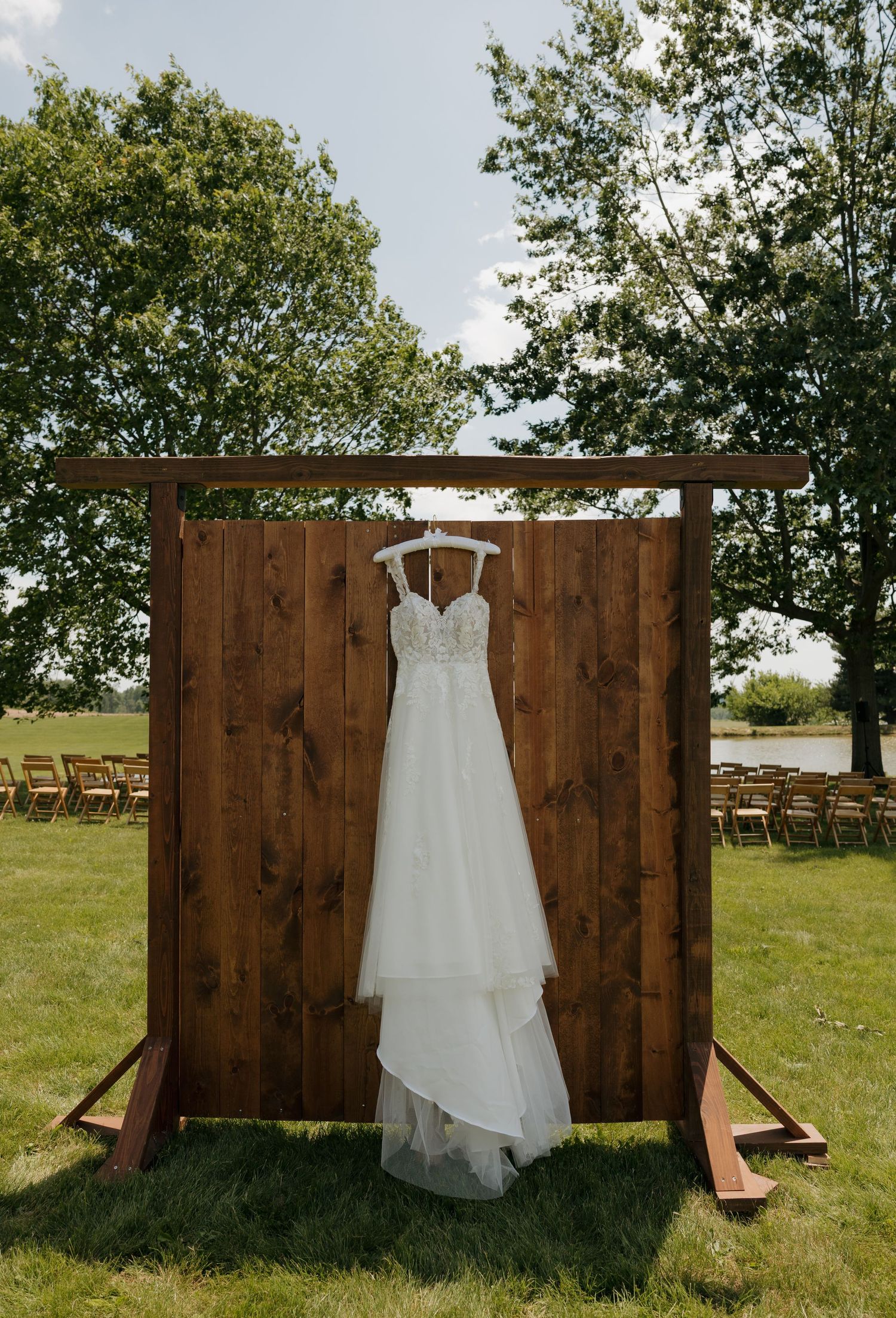 White bridal gown hanging on rustic wooden backdrop outdoors, with ceremony chairs and lush green trees in background.