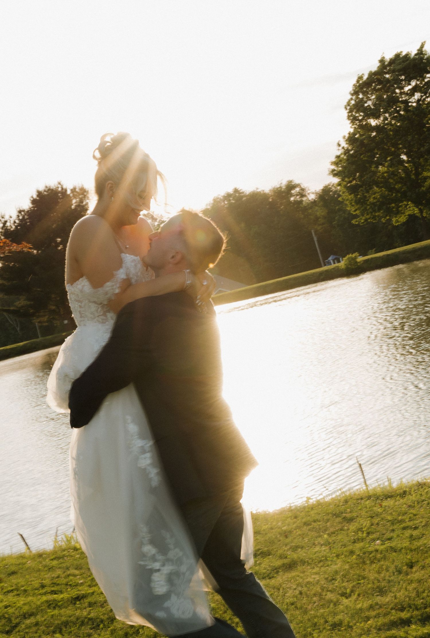 Groom lifting bride in wedding dress by a sunlit lake, sharing a romantic moment at golden hour.