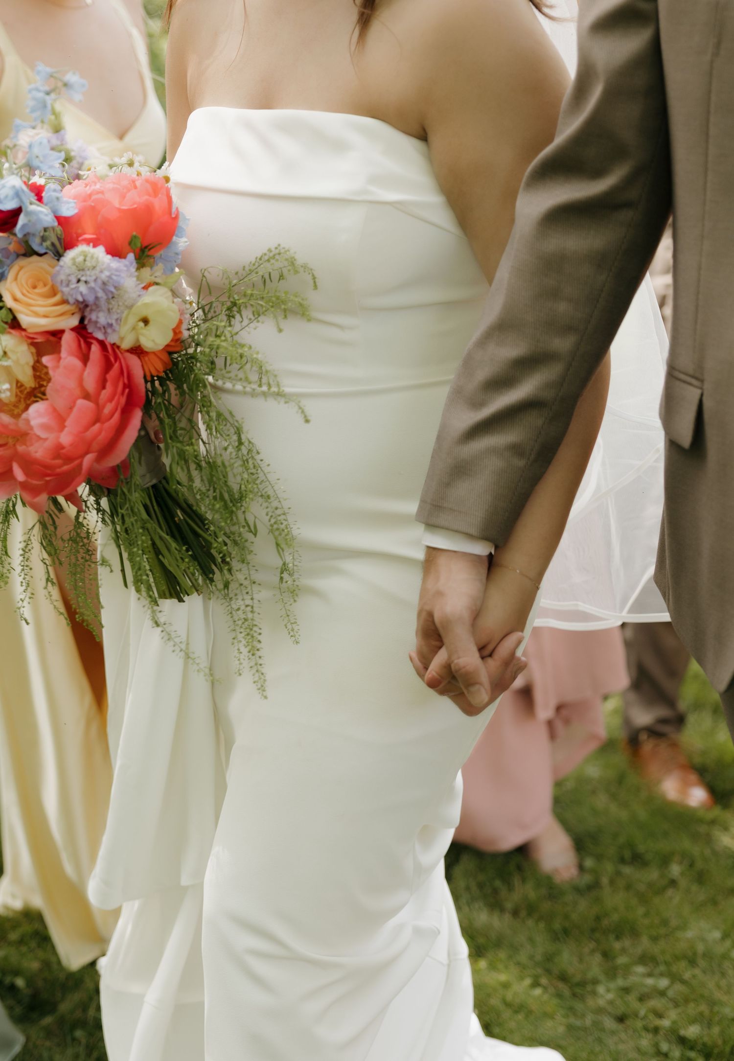 Bride in white strapless gown holding colorful bridal bouquet with peonies, groom in tan suit holding hands outdoors.