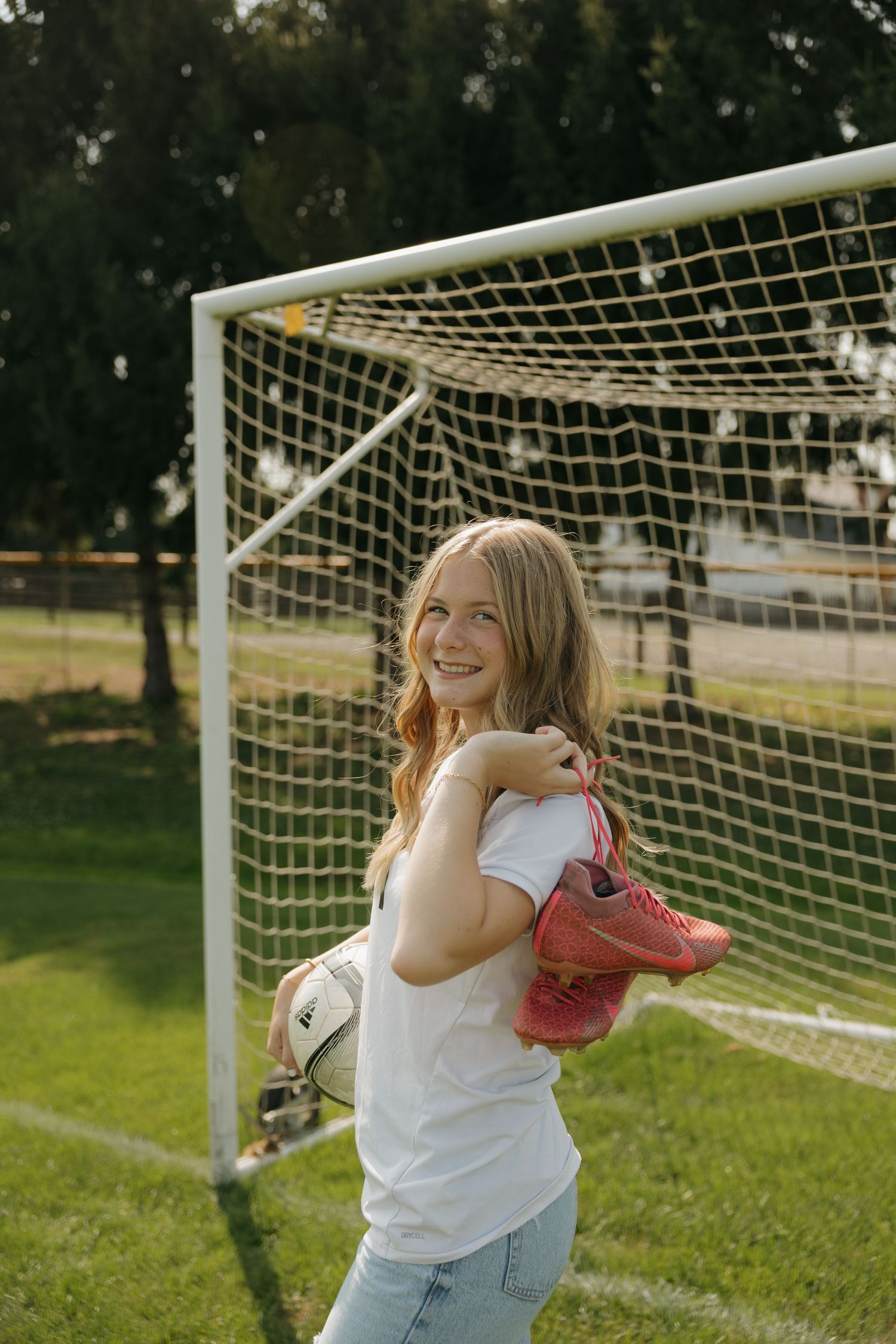 Smiling teenage girl holding soccer ball and pink cleats standing in front of a white goal net on a green field.
