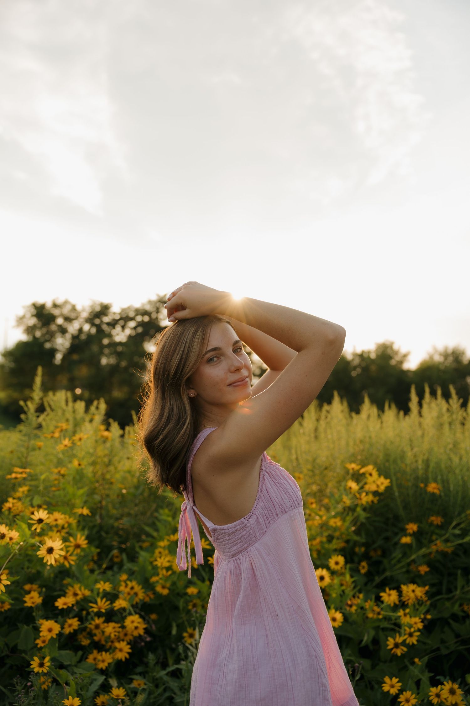 Young woman in pink dress posing among yellow wildflowers at golden hour sunset in a lush green field.