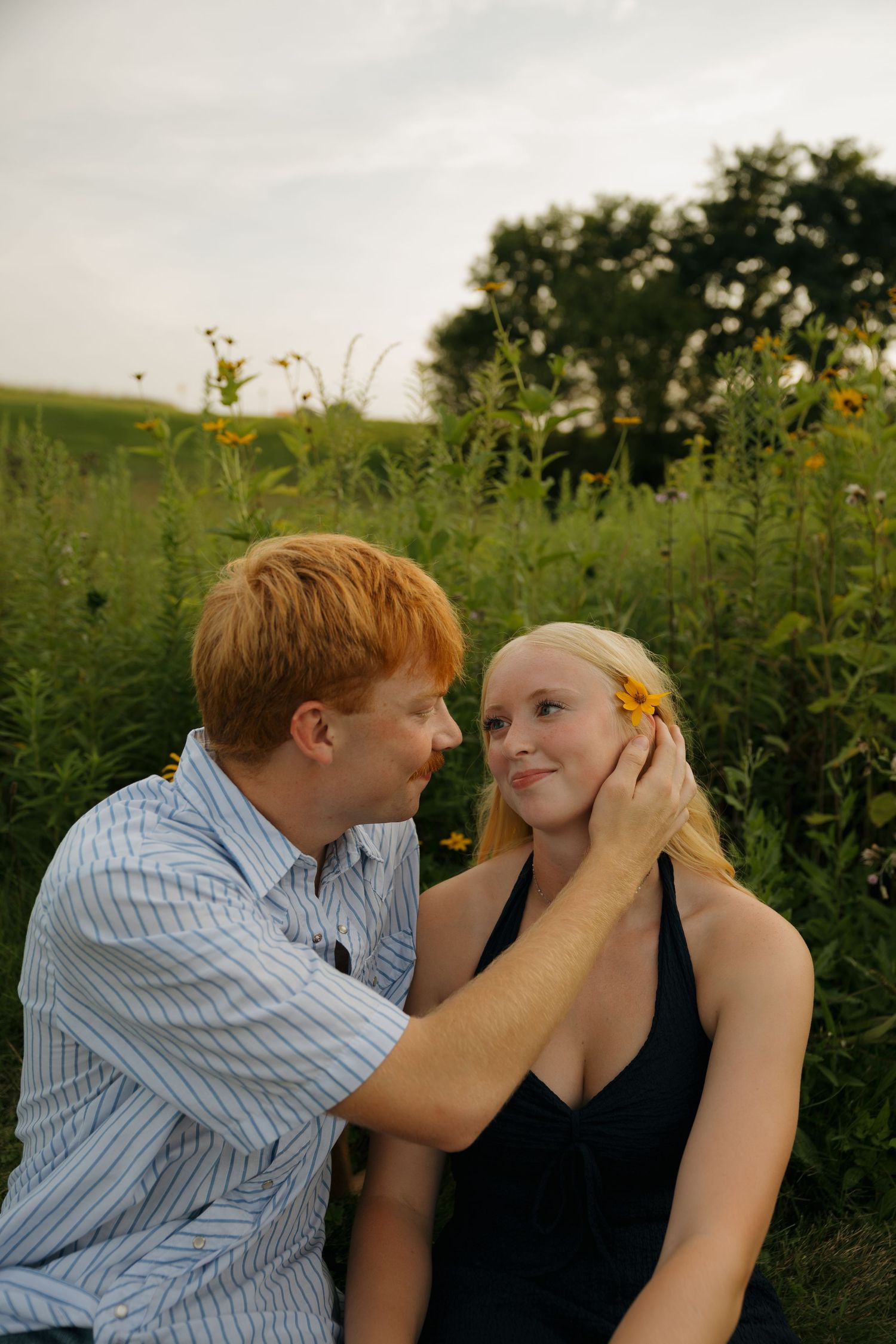 A young couple sits in a wildflower field, as a red-haired man gently places a yellow flower in a blonde woman's hair.