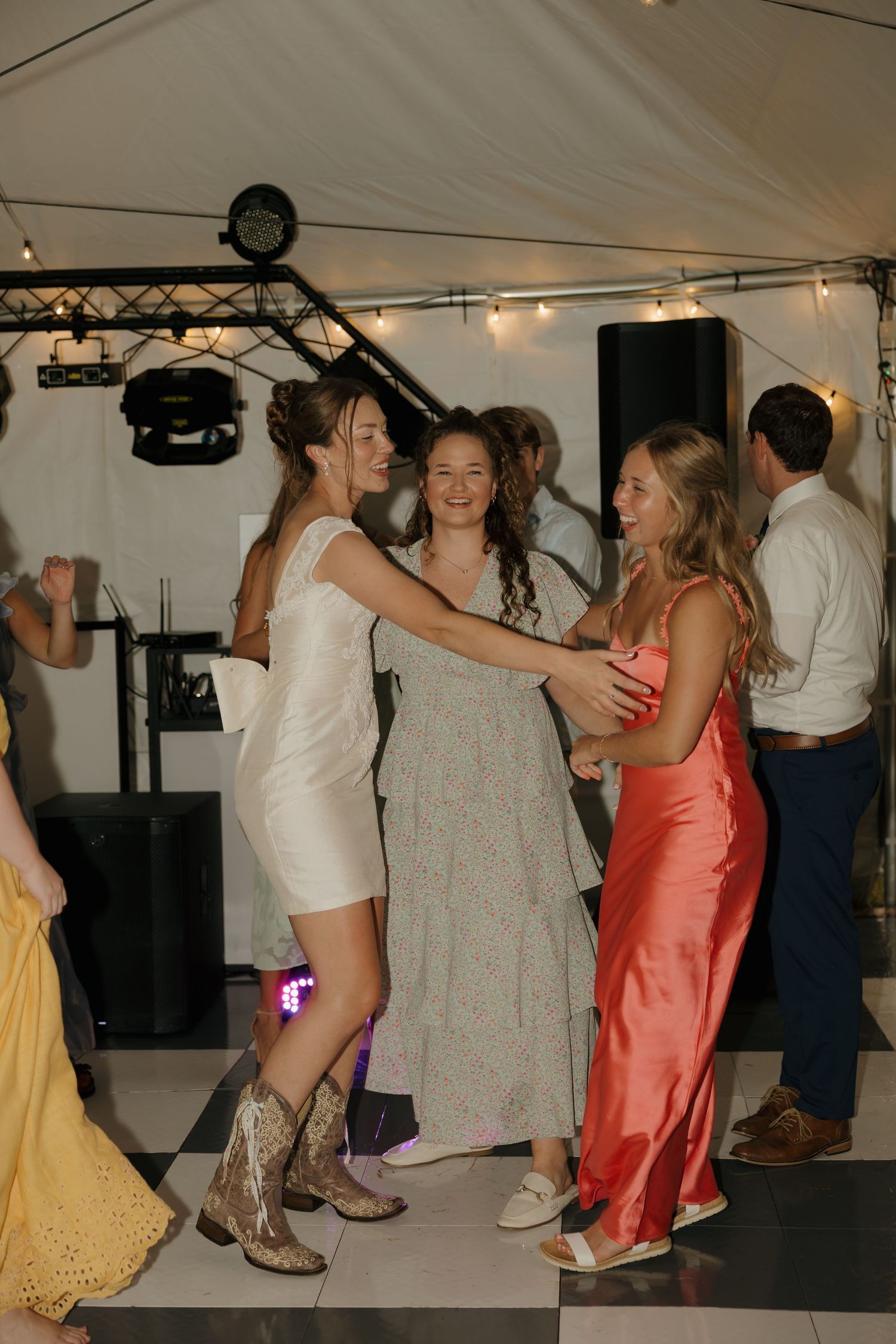 Women in elegant dresses dancing together at an outdoor wedding reception under a white tent with string lights.