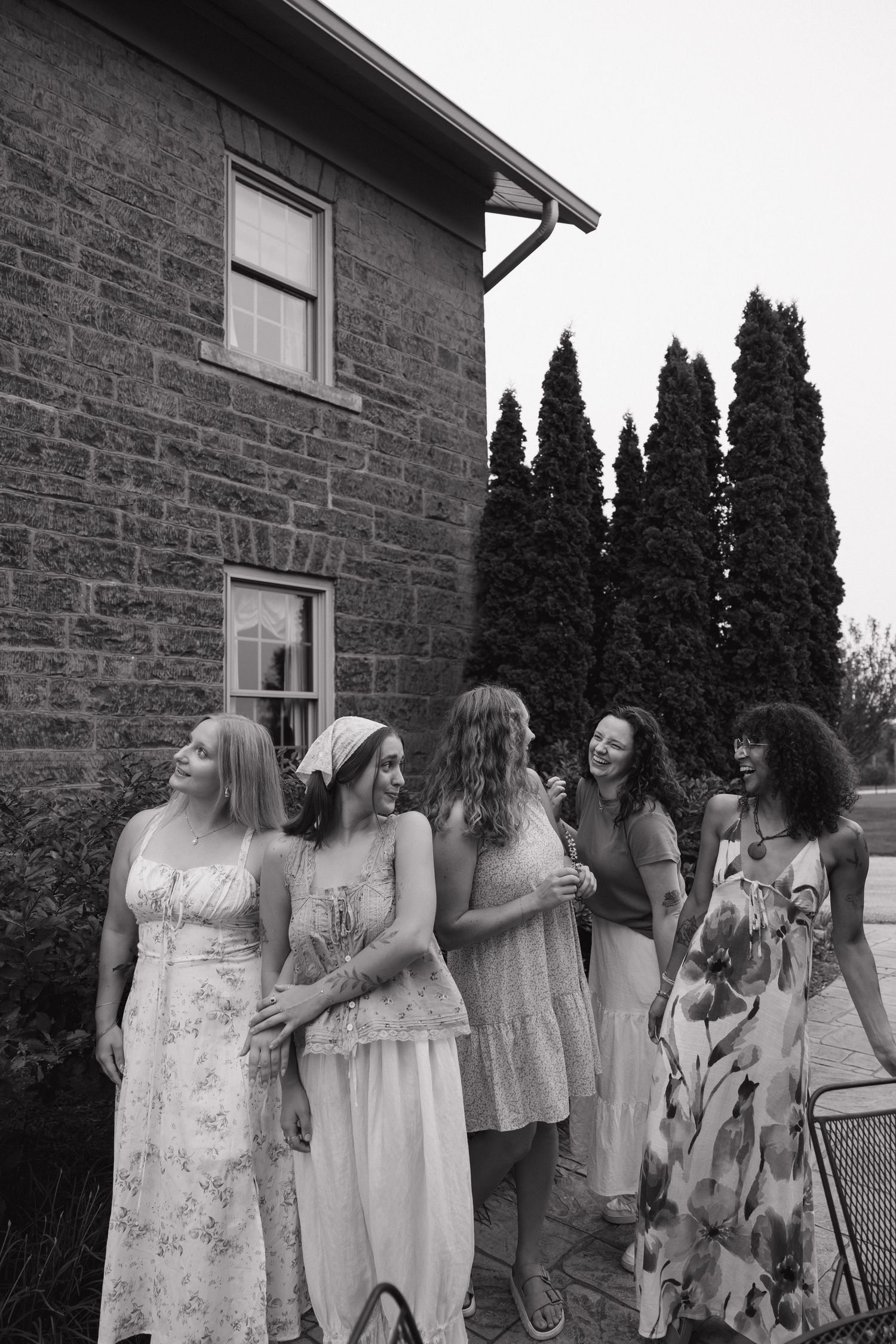 Five women in summer dresses laugh together outside historic stone building with tall cypress trees.