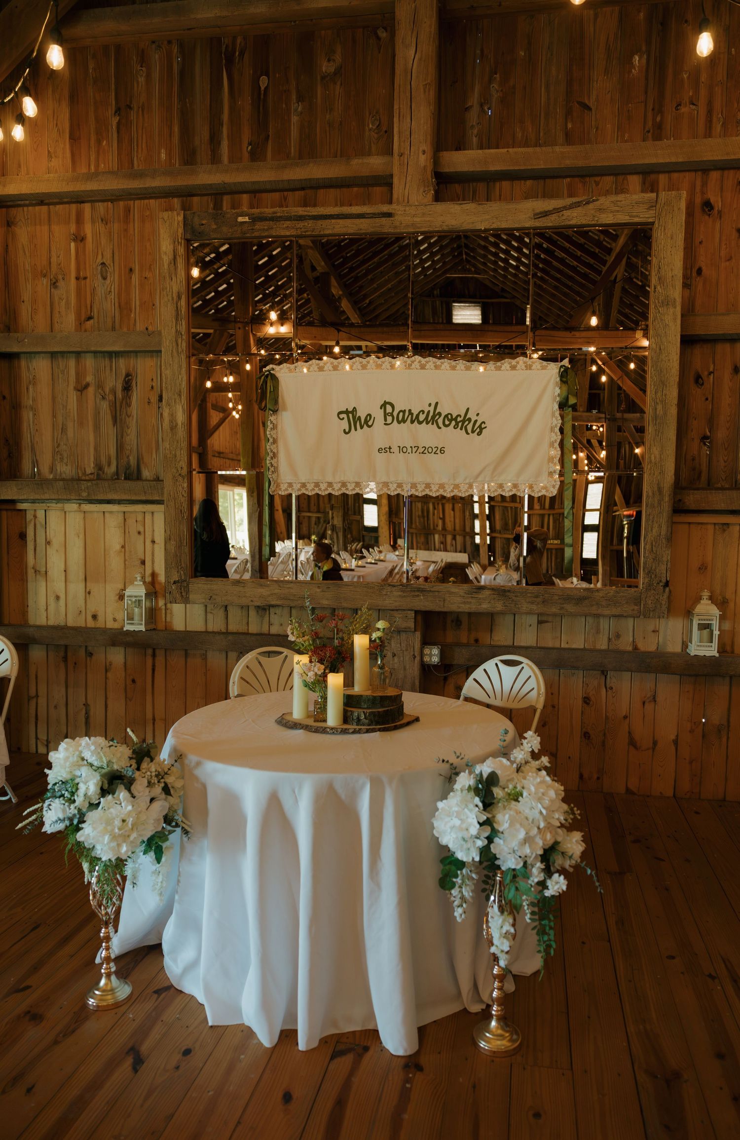 Rustic barn wedding sweetheart table with white linen, candles and white floral arrangements beneath a hanging banner and string lights.