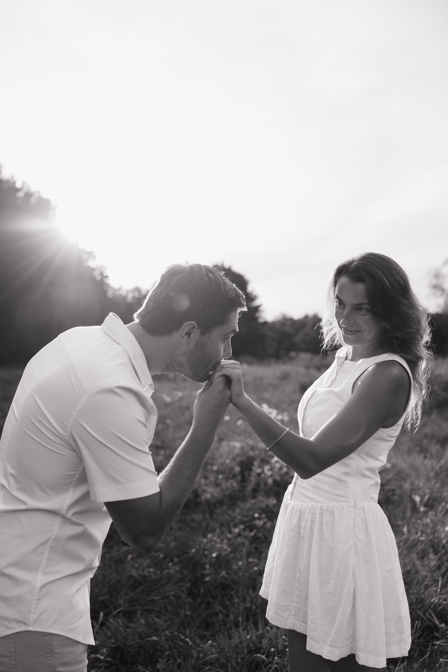 Black and white photo of a man kissing a woman's hand in a sunlit field, romantic outdoor couple portrait.