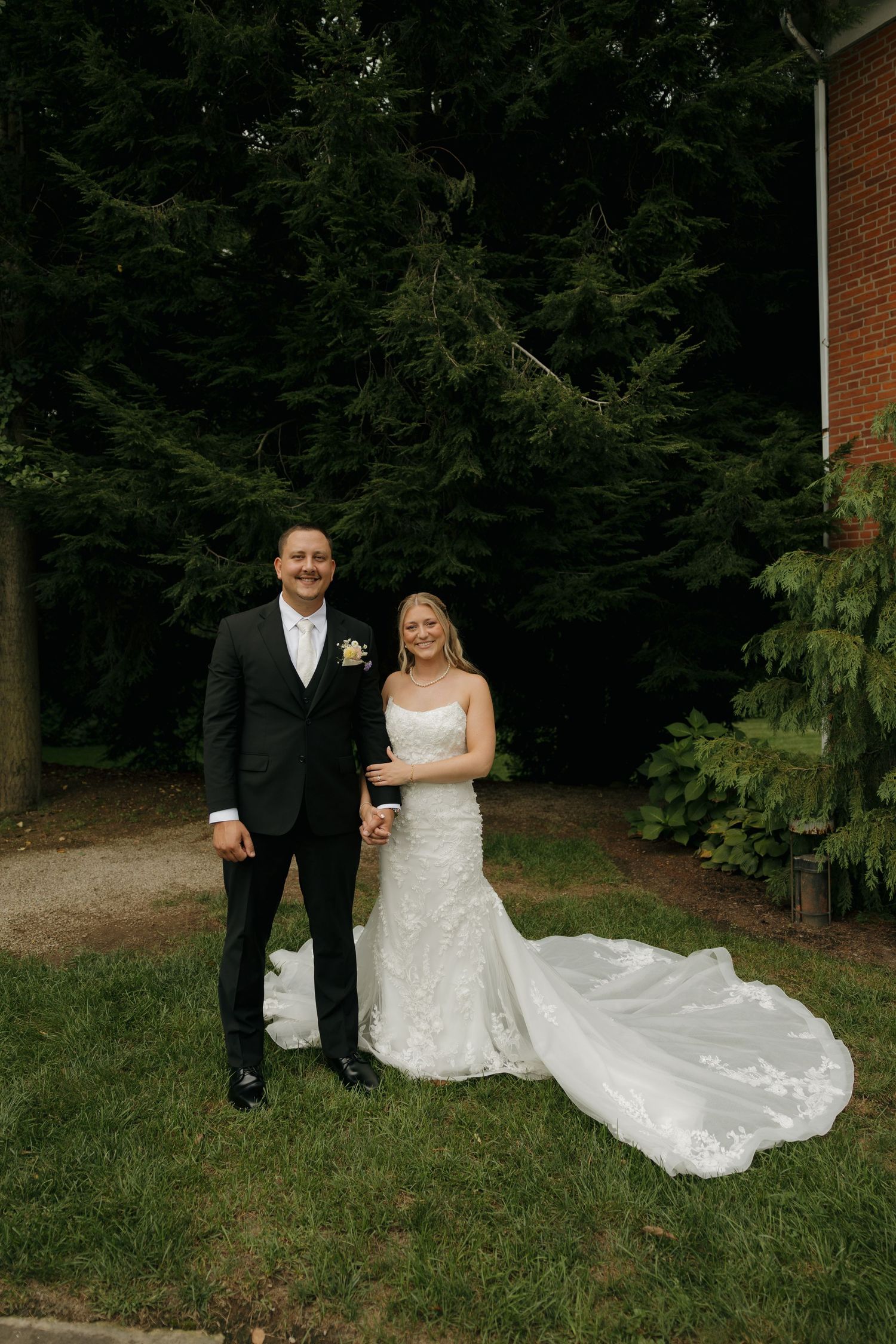 Bride and groom smiling together outdoors on their wedding day, with lush green trees and a brick building in the background.