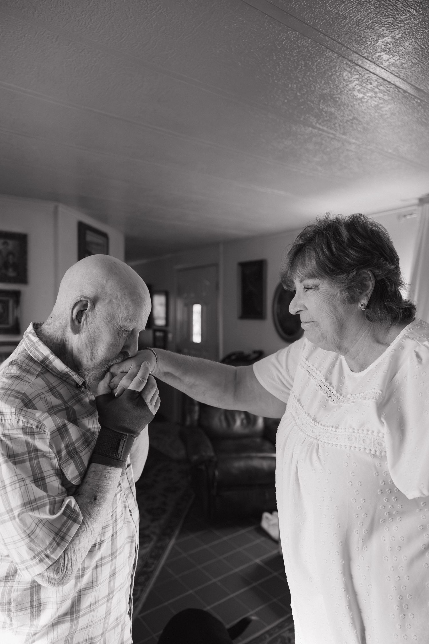 Elderly man kissing a woman's hand in black and white photo, sharing a tender romantic moment indoors.