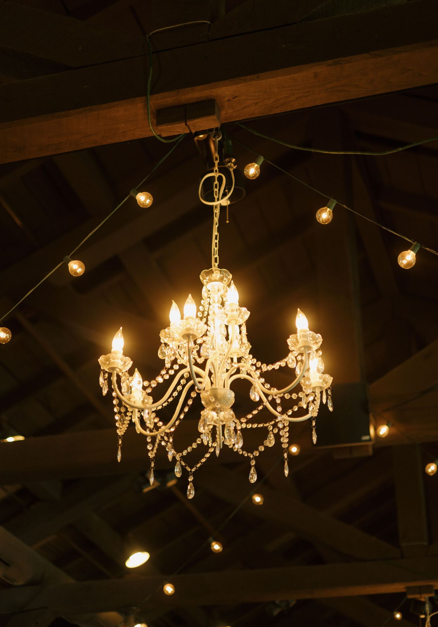 Elegant crystal chandelier with glowing candle lights hanging from rustic wooden ceiling beams in a warm venue.