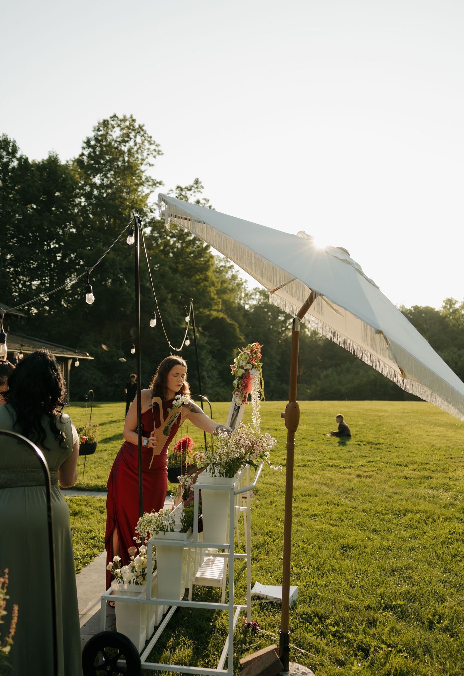 Woman in red dress serving drinks from white cart under fringed umbrella at outdoor wedding reception.