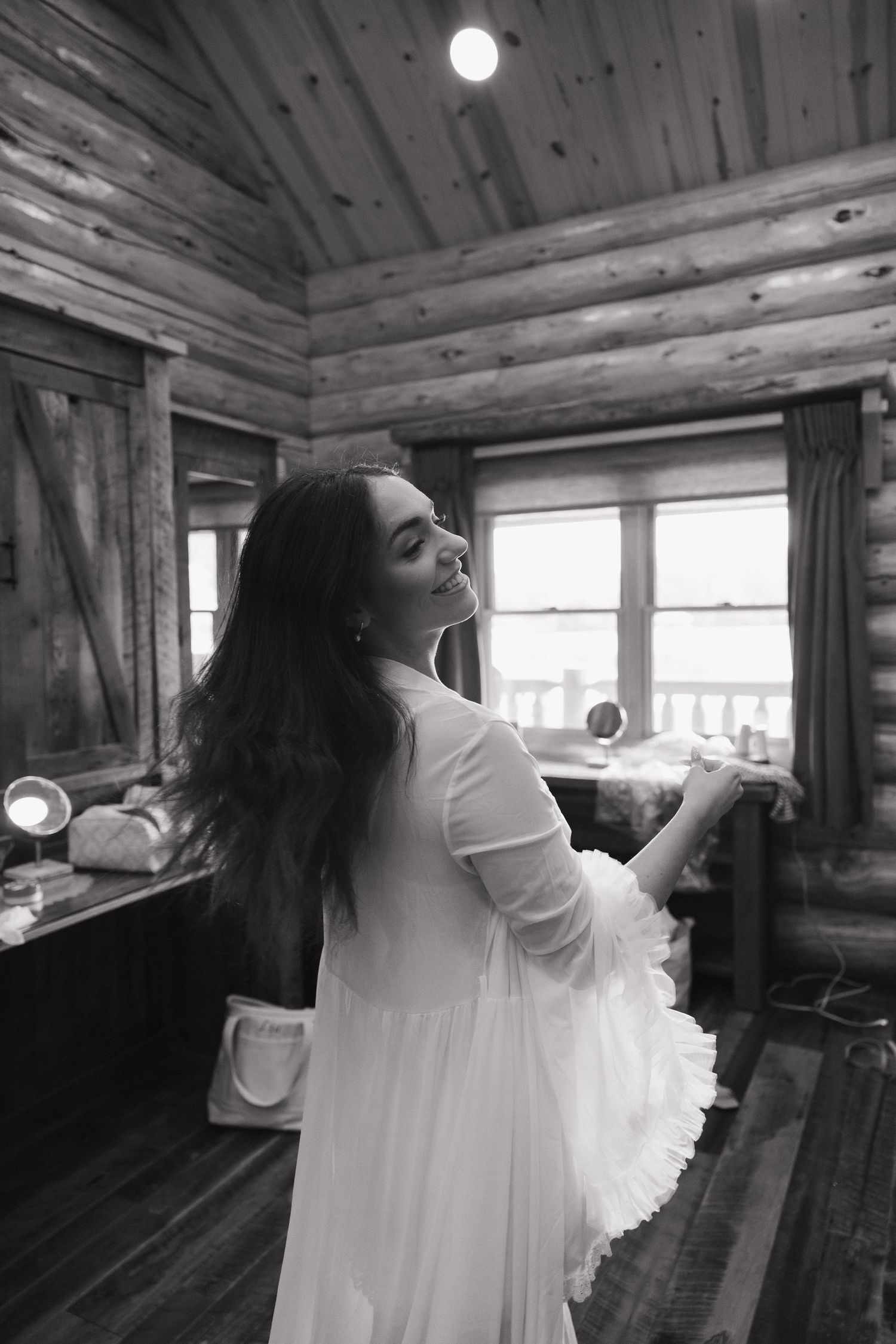 Black and white photo of a woman in a white dress smiling in a rustic log cabin interior.