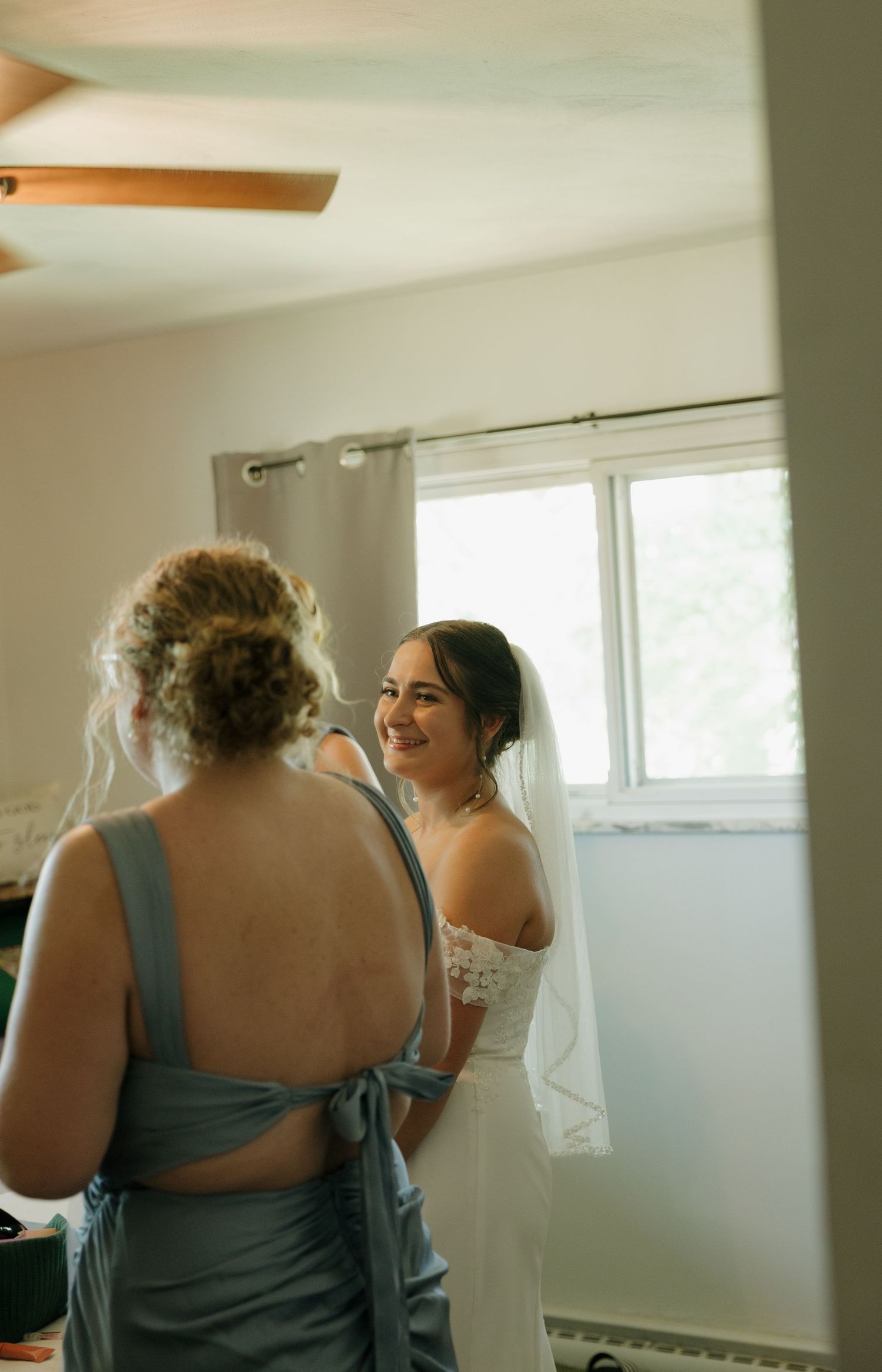 Smiling bride in off-shoulder wedding dress and veil gets ready with bridesmaid in blue dress before ceremony.