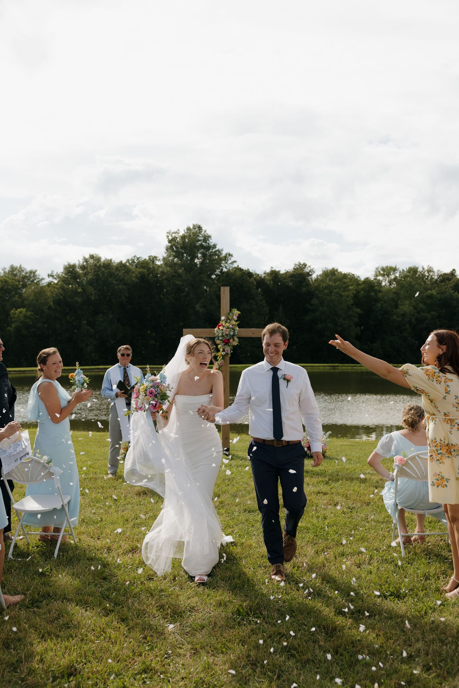 Bride and groom walk down outdoor aisle as guests toss white flower petals near a lake.