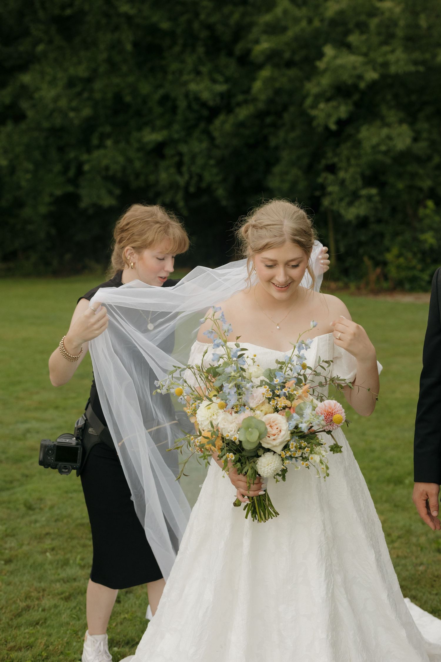 Bride in white gown holds floral bouquet while assistant adjusts veil outdoors on green lawn.