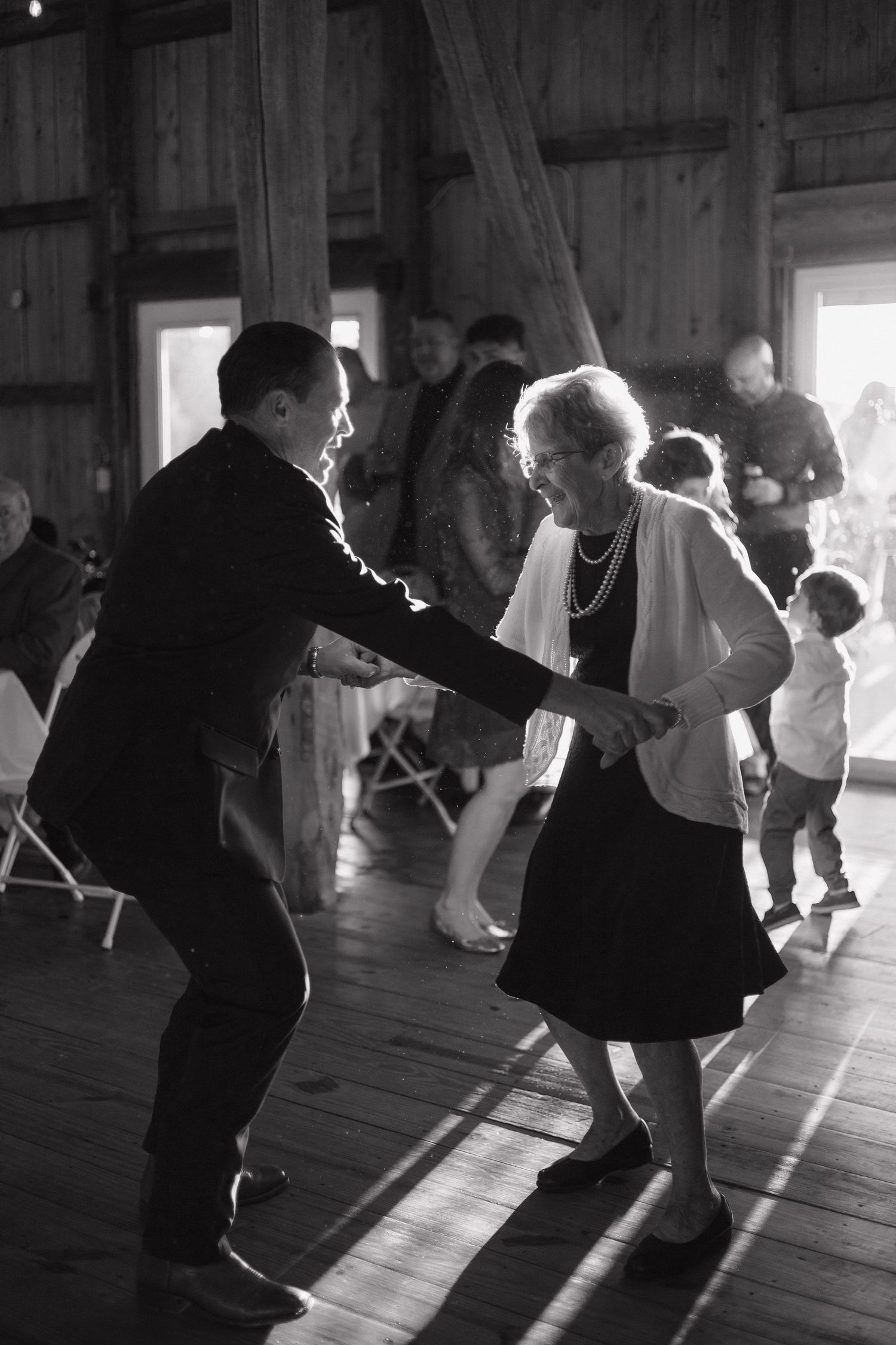 A man in a dark suit dances joyfully with a smiling older woman in a cardigan on a wooden barn floor during a wedding reception.