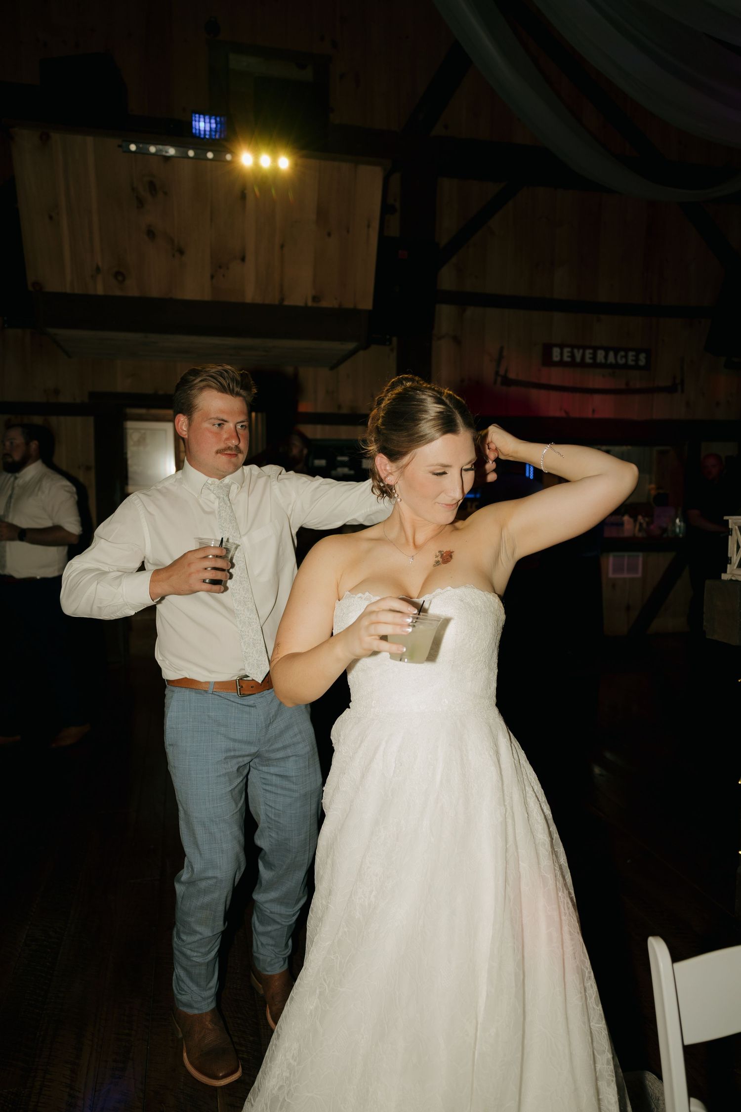 A bride in a strapless white gown dances with a drink in hand beside a man in a white shirt inside a rustic wooden barn venue.