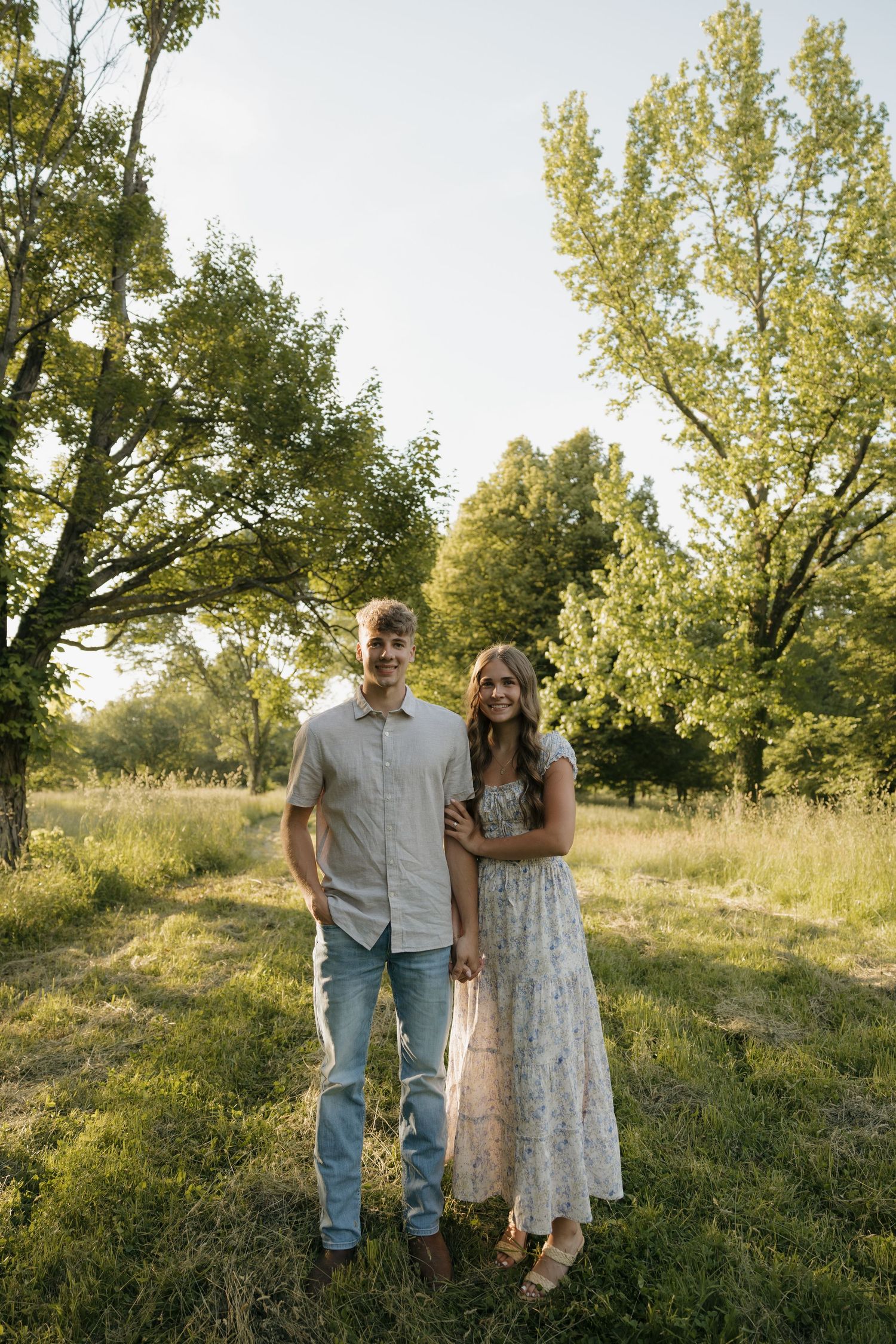 A young couple stands together in a lush green field, smiling, with tall trees in the background during golden hour.