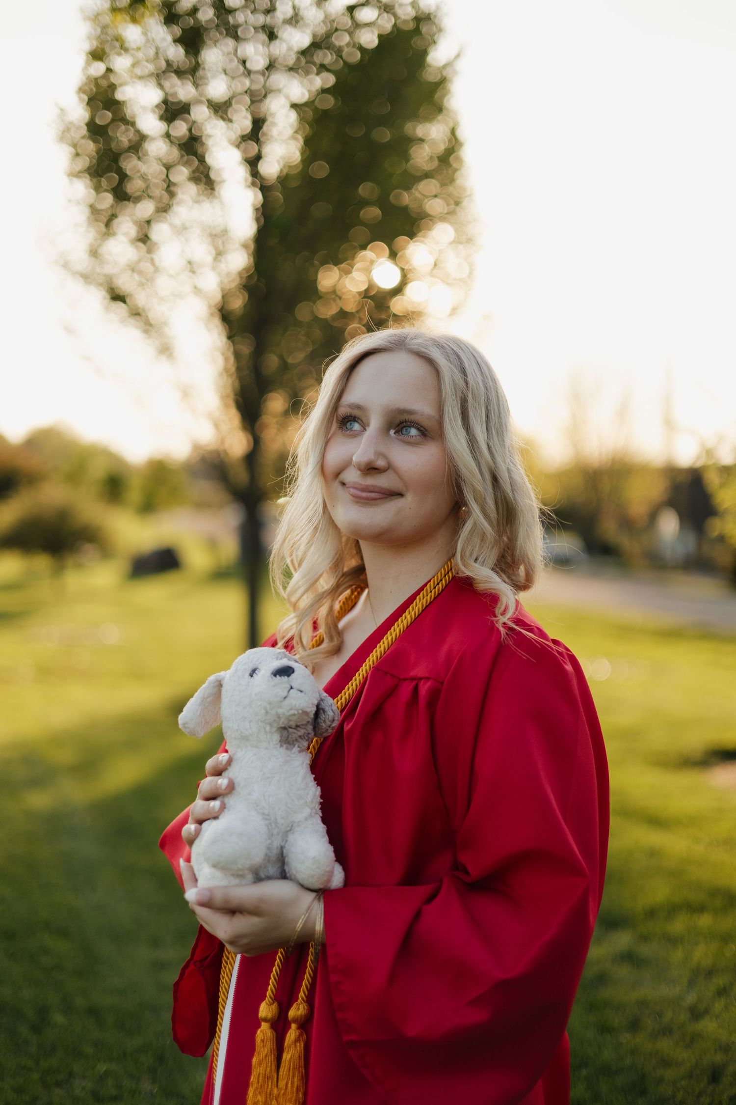 Young woman in red graduation gown holding a white stuffed animal outdoors on a sunny day.
