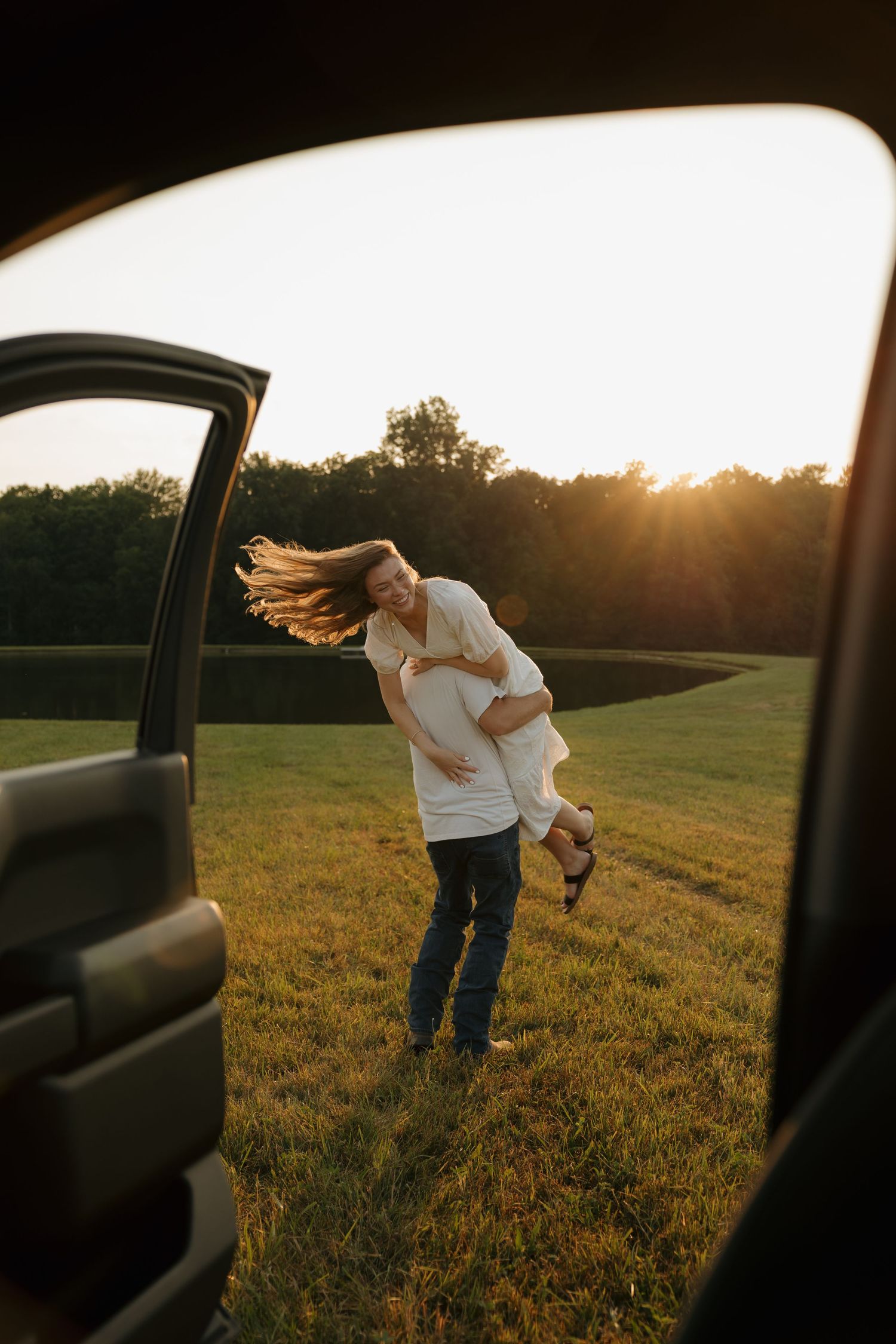 Couple embracing at golden sunset in green field, viewed through open car door, woman's hair blowing in wind.