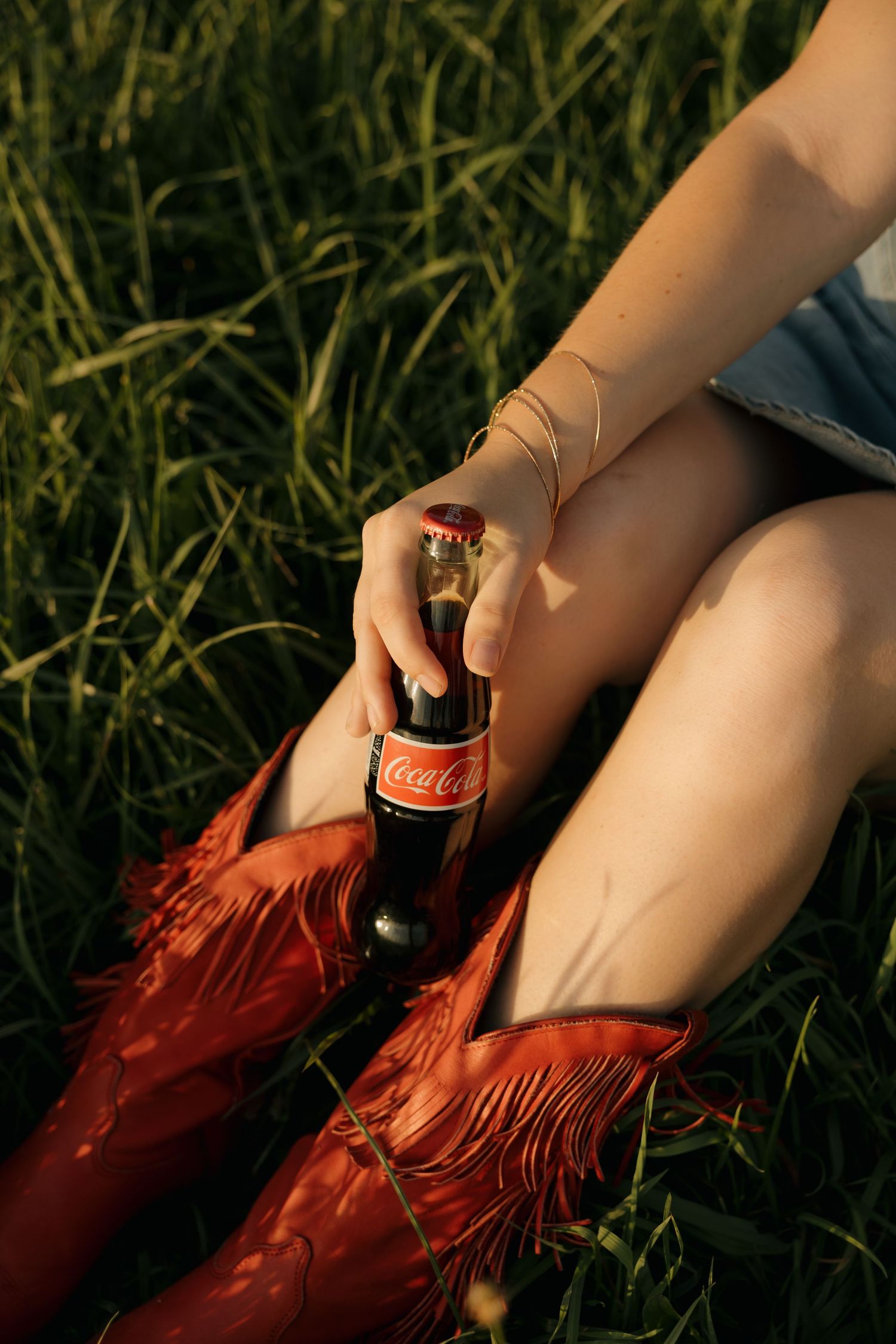 Woman sitting in grass holding a Coca-Cola glass bottle, wearing red fringe cowboy boots, golden hour lighting.