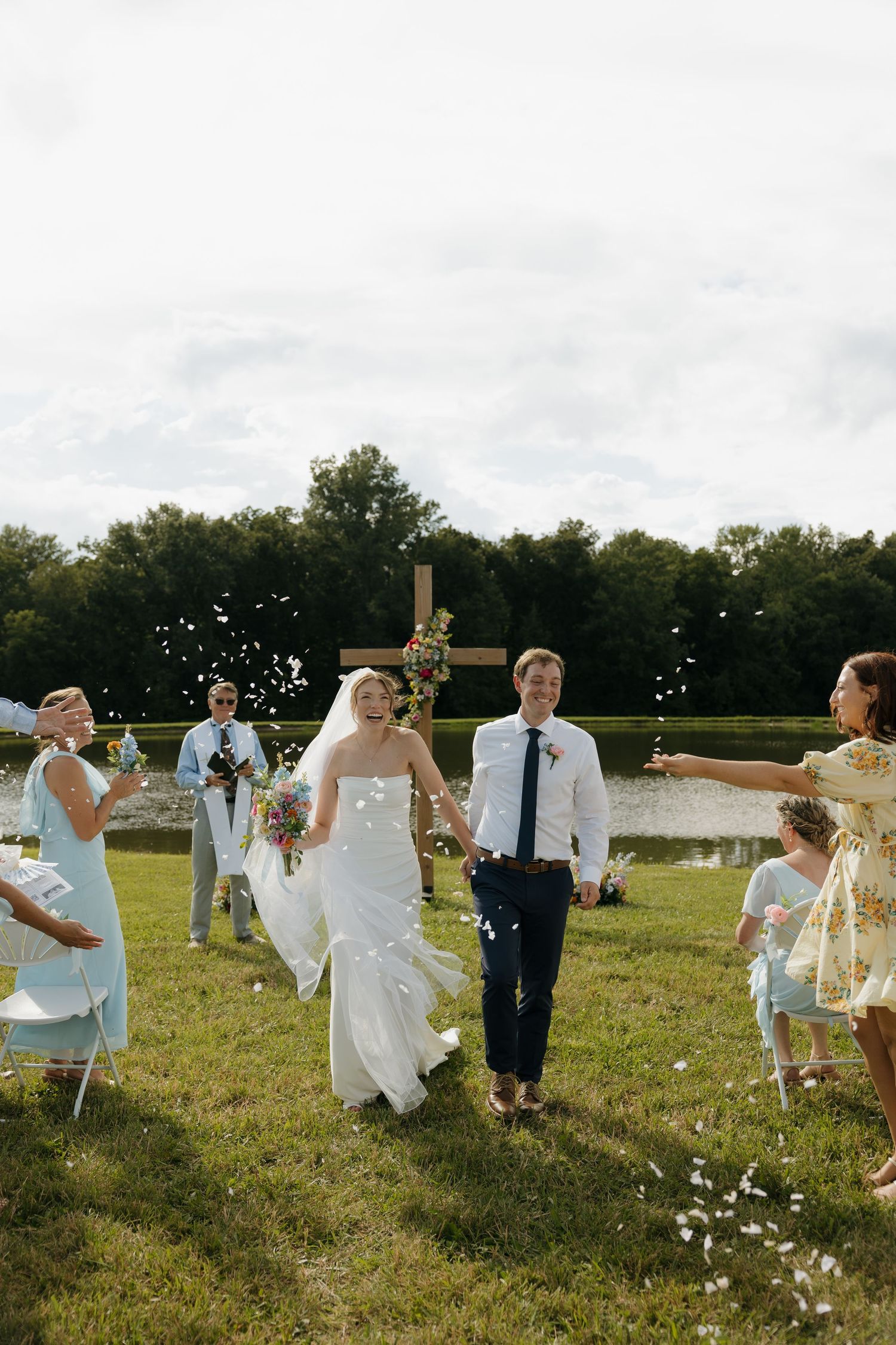 Bride and groom walk down outdoor aisle as guests toss flower petals, with wooden cross and pond in background.