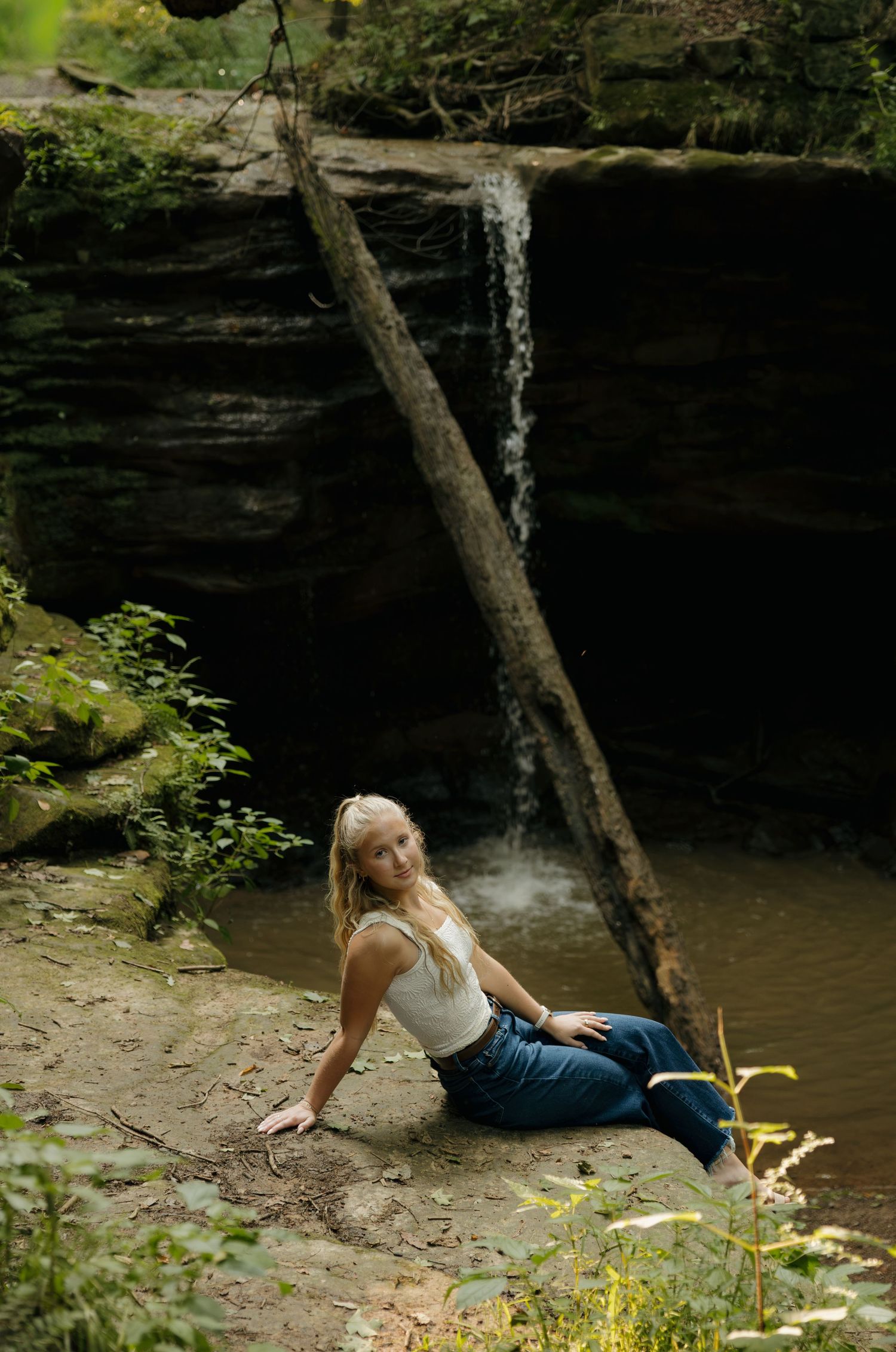 Blonde woman in white top and jeans posing by a small waterfall and rocky creek in a lush forest setting.