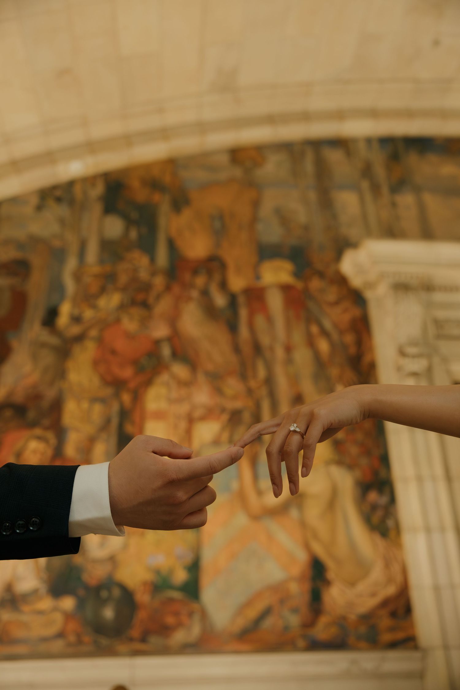 Newlywed couple touching fingers with wedding ring visible, elegant tapestry mural backdrop in ornate venue.