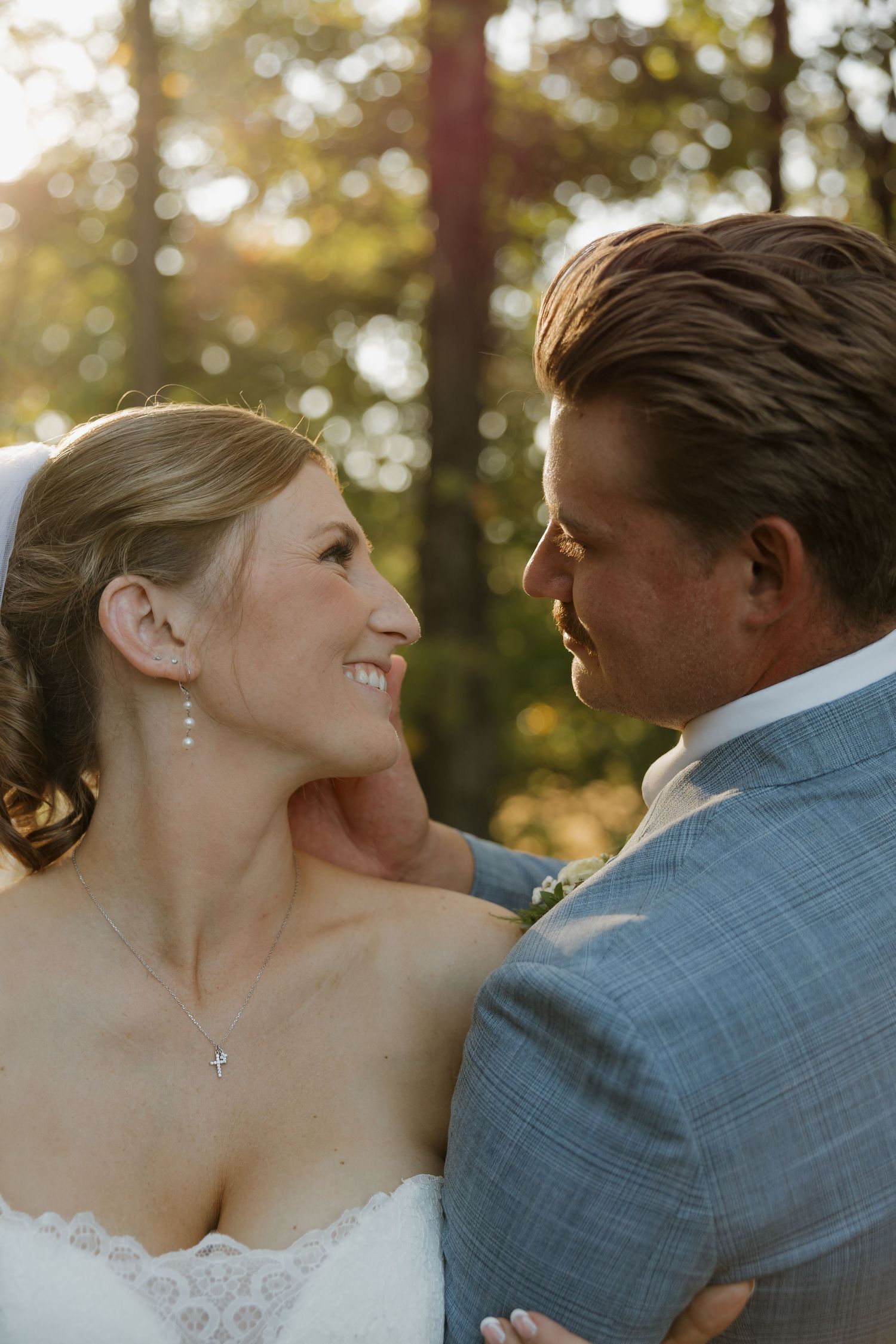 Smiling bride in a lace gown and pearl earrings embraces her groom in a gray-blue suit during a sunlit outdoor wedding portrait among trees.