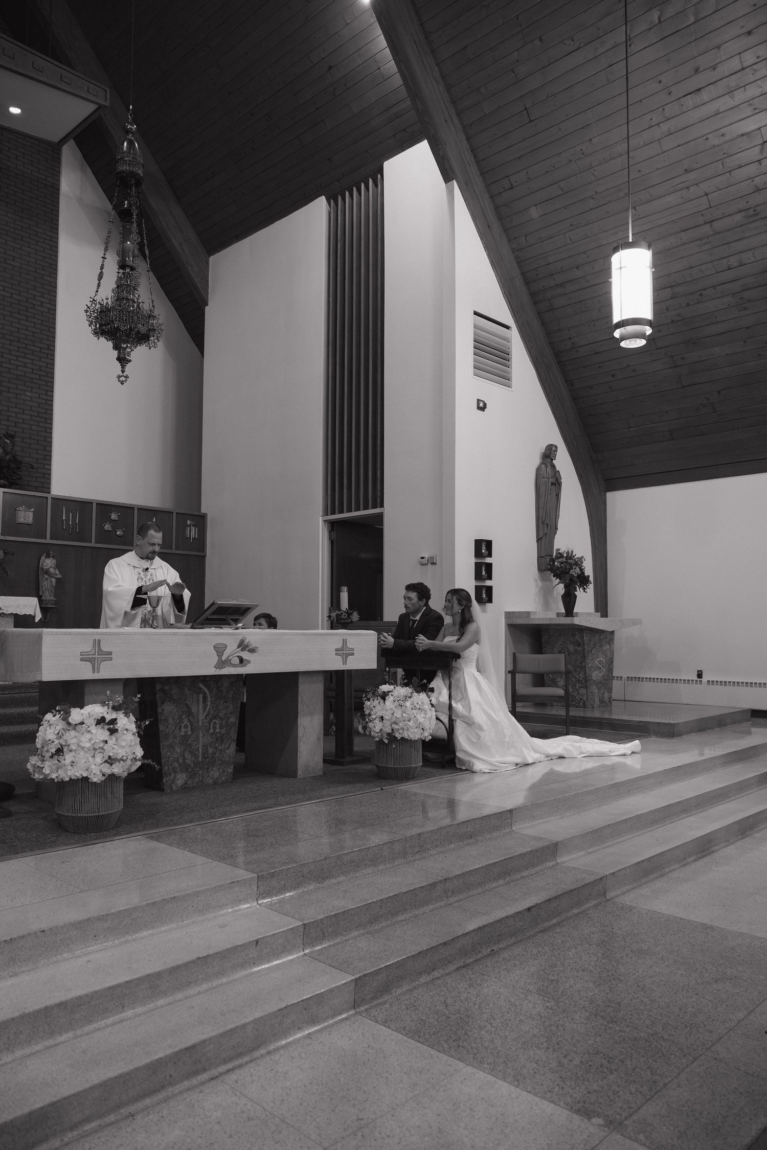 Black and white photo of a bride and groom kneeling at a church altar during a wedding ceremony as a priest officiates.