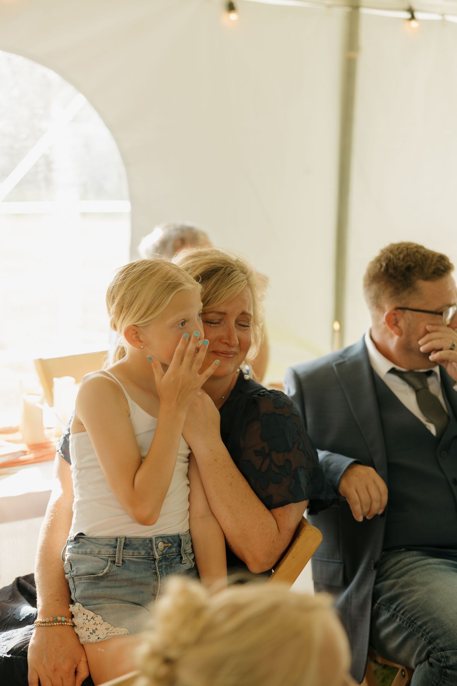 Emotional wedding guest wipes tears as young girl comforts her during outdoor tent ceremony.
