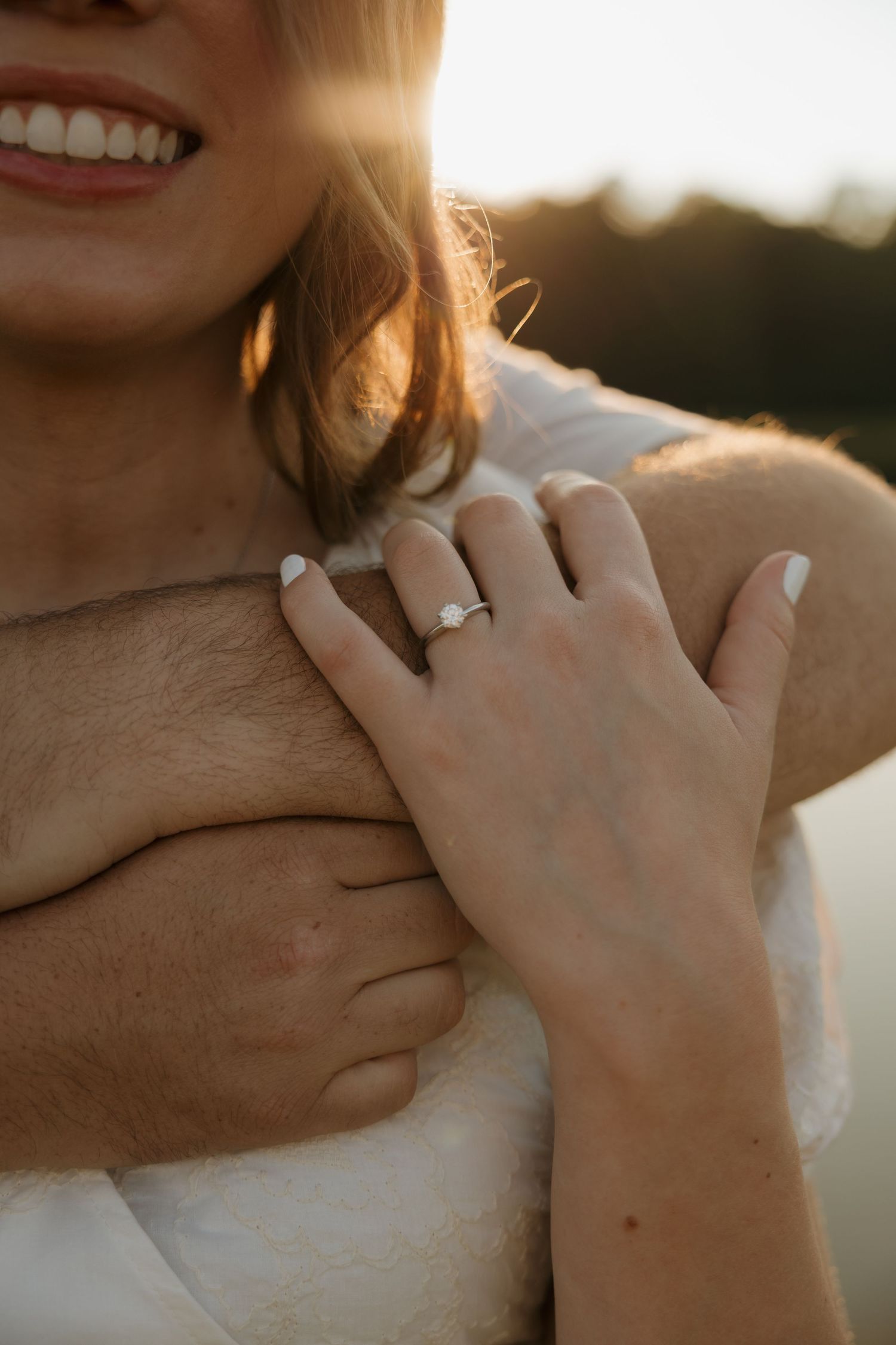 Close-up of a smiling bride resting her hand on groom's chest, showcasing a diamond engagement ring at golden hour.