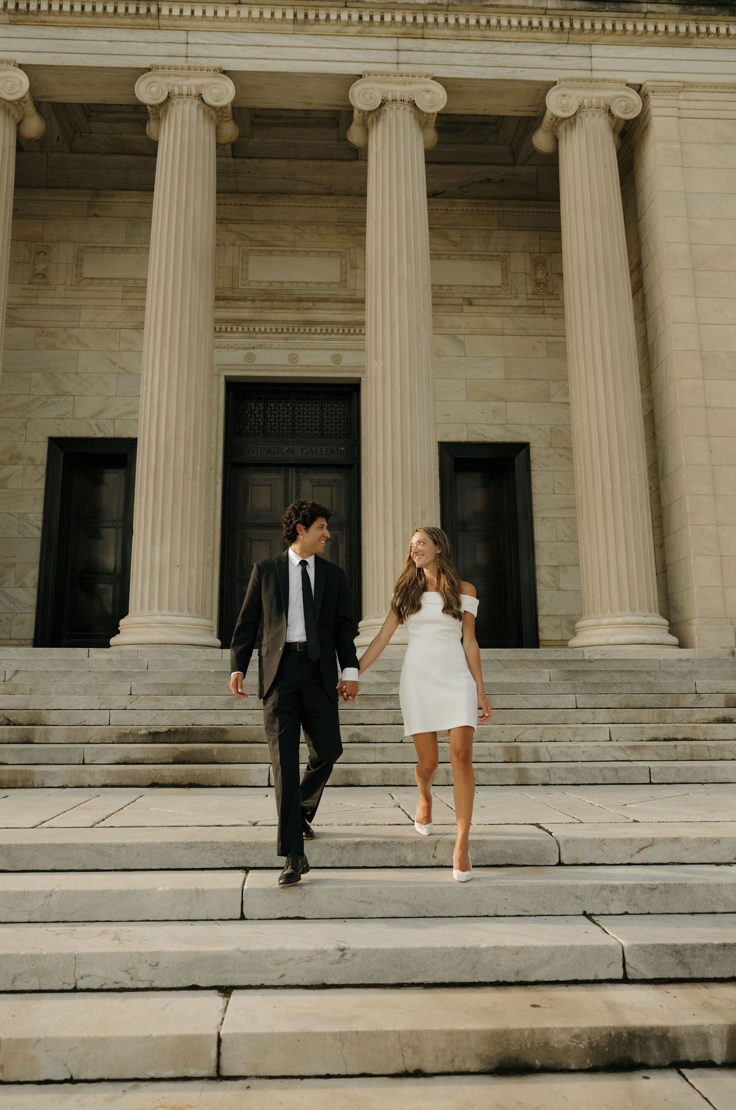 Couple in wedding attire walking down steps of a grand columned building, bride in short white dress.