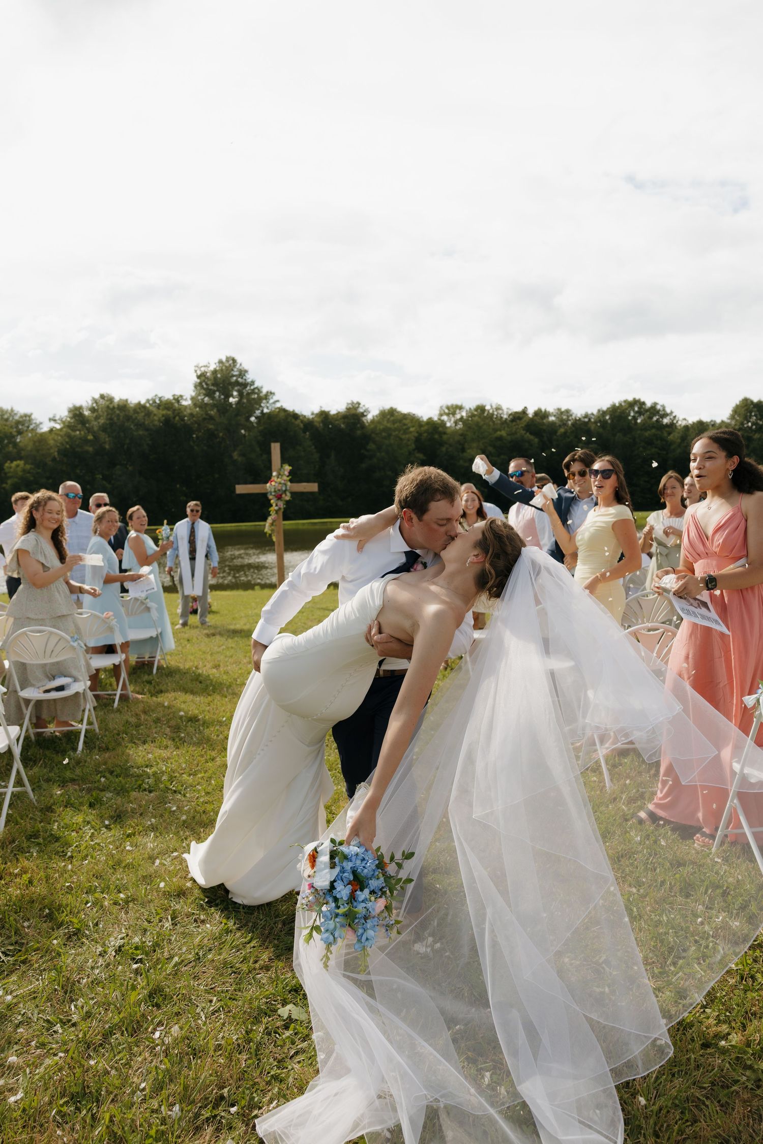 Bride and groom share a romantic dip kiss at outdoor wedding ceremony with guests cheering on green lawn.