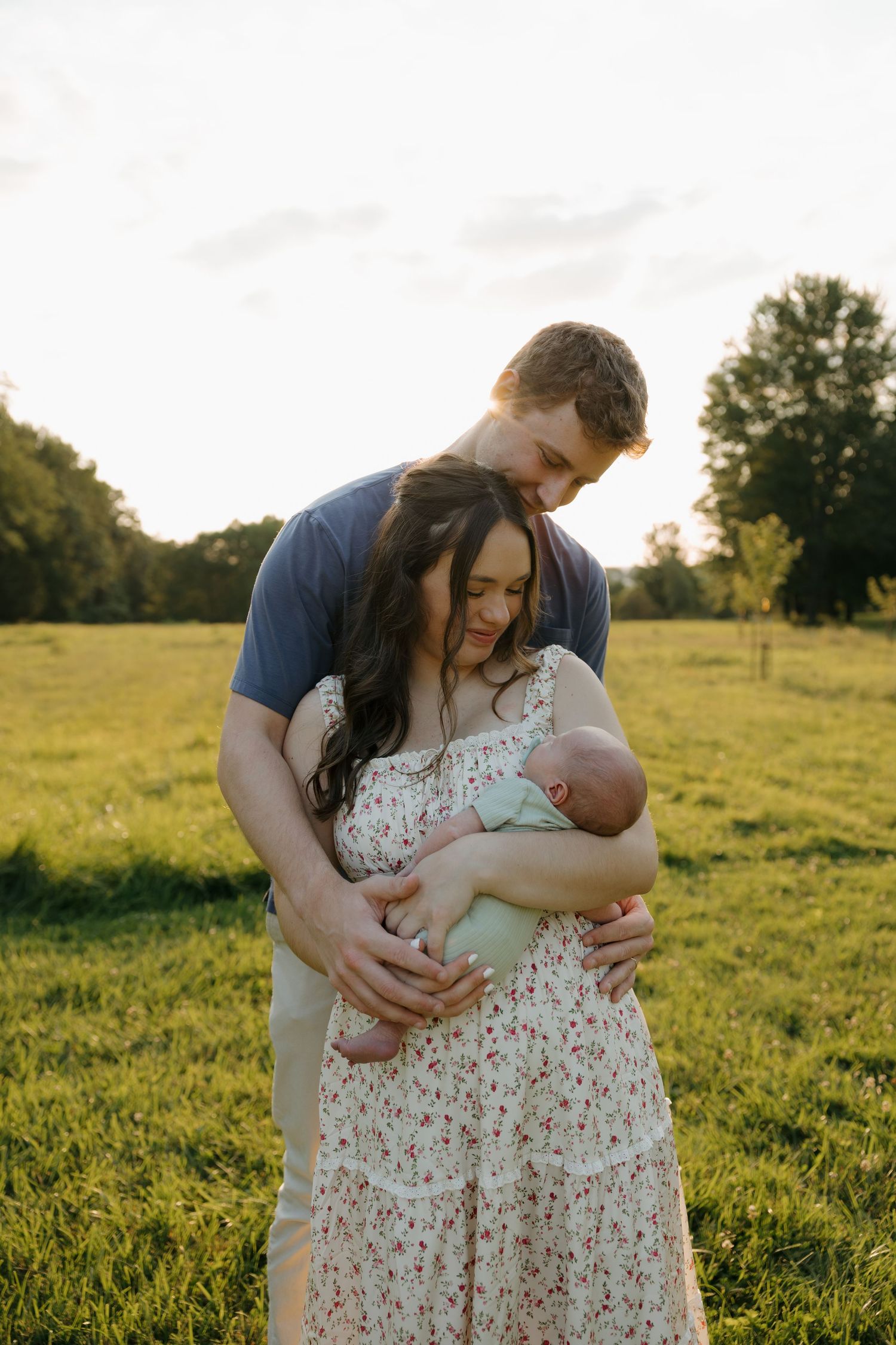 Young couple embracing outdoors at golden hour, tenderly cradling their newborn baby in a sunlit green meadow.