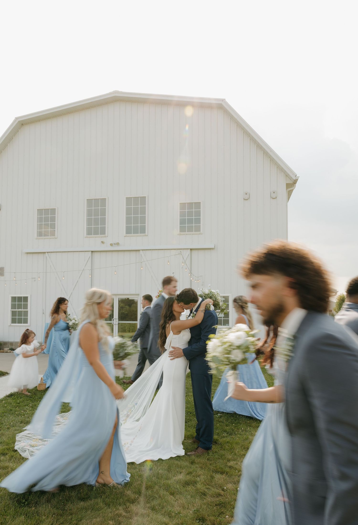 Wedding party celebrates outside a white barn as bride and groom share a romantic kiss.