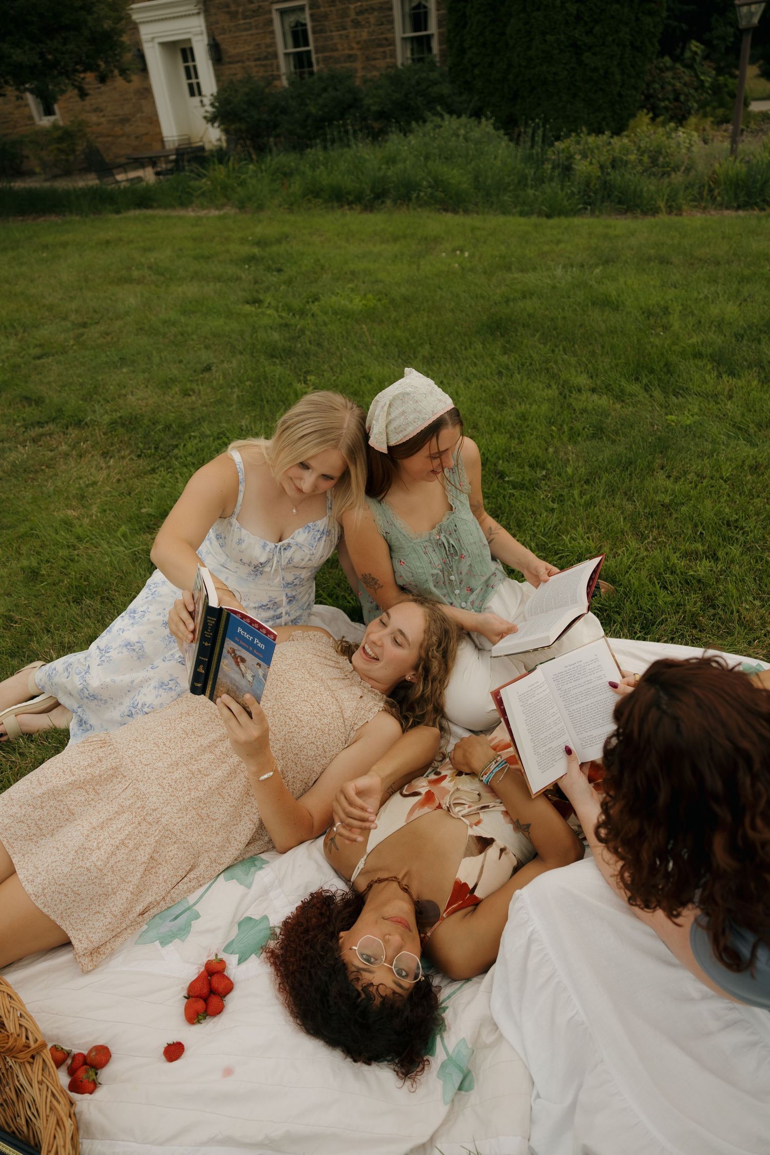 Four women enjoy a summer picnic on a blanket, reading books and sharing strawberries on a green lawn.