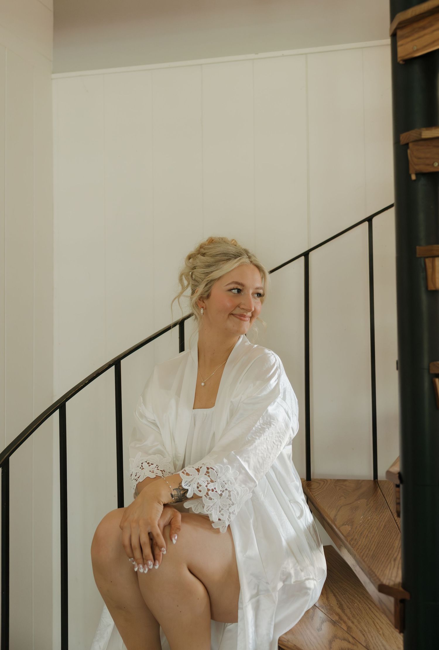Smiling bride in white lace robe sitting on wooden spiral staircase, looking to the side on her wedding day.