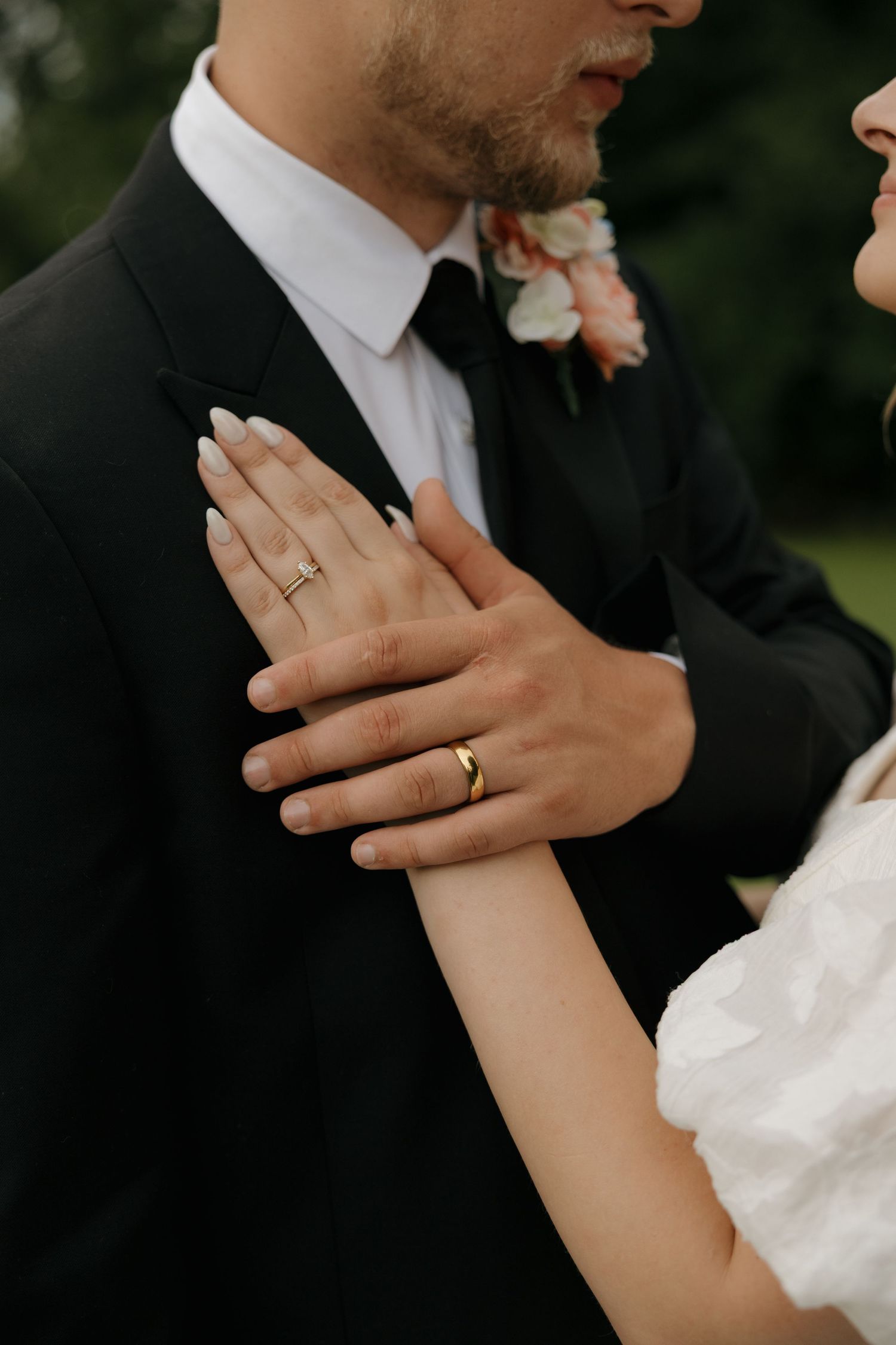 Close-up of bride and groom hands showing gold wedding rings, bride in white dress touching groom in black suit.