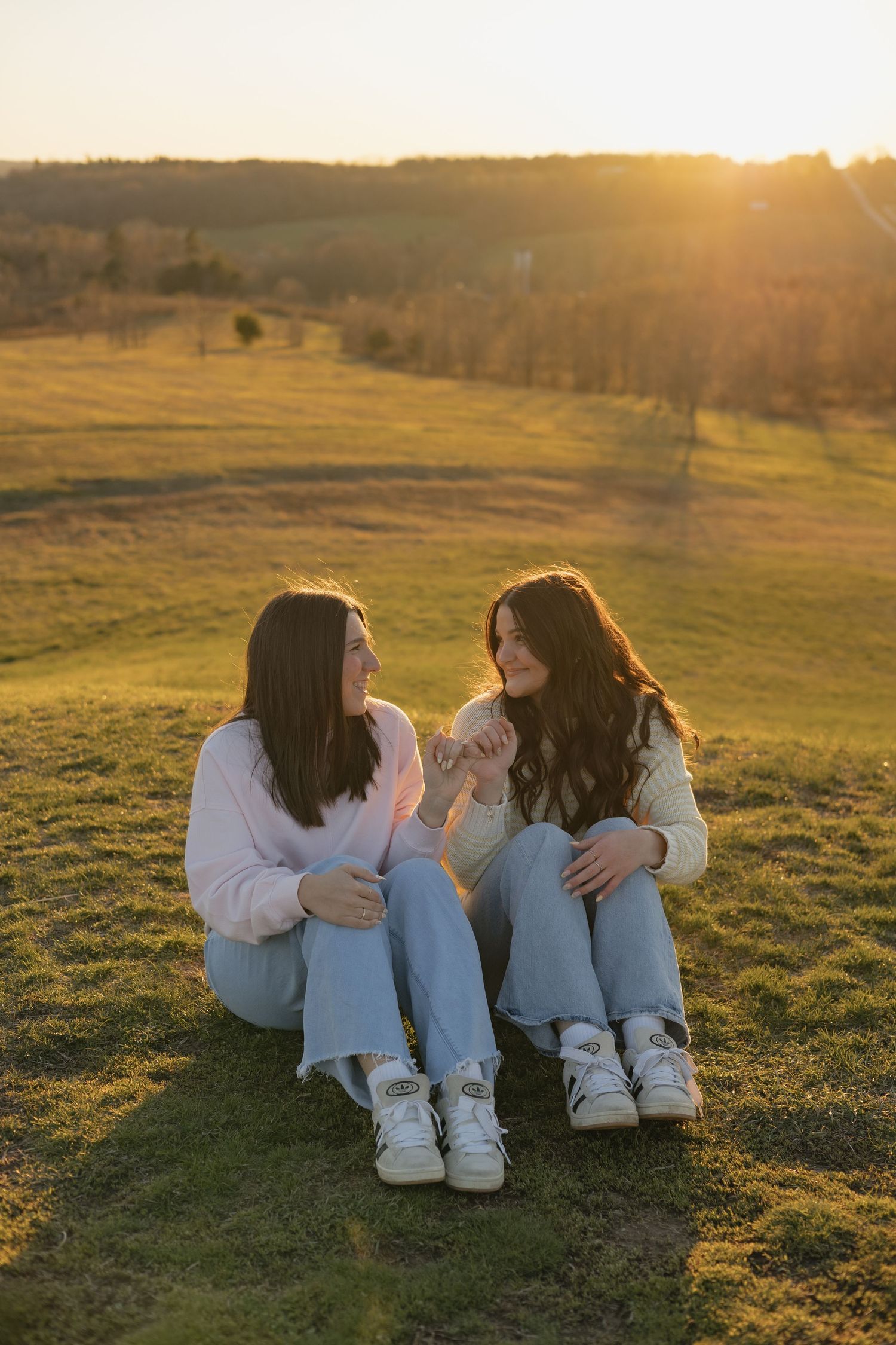 Two people in white sweaters and jeans sit together on grassy hill during golden sunset.