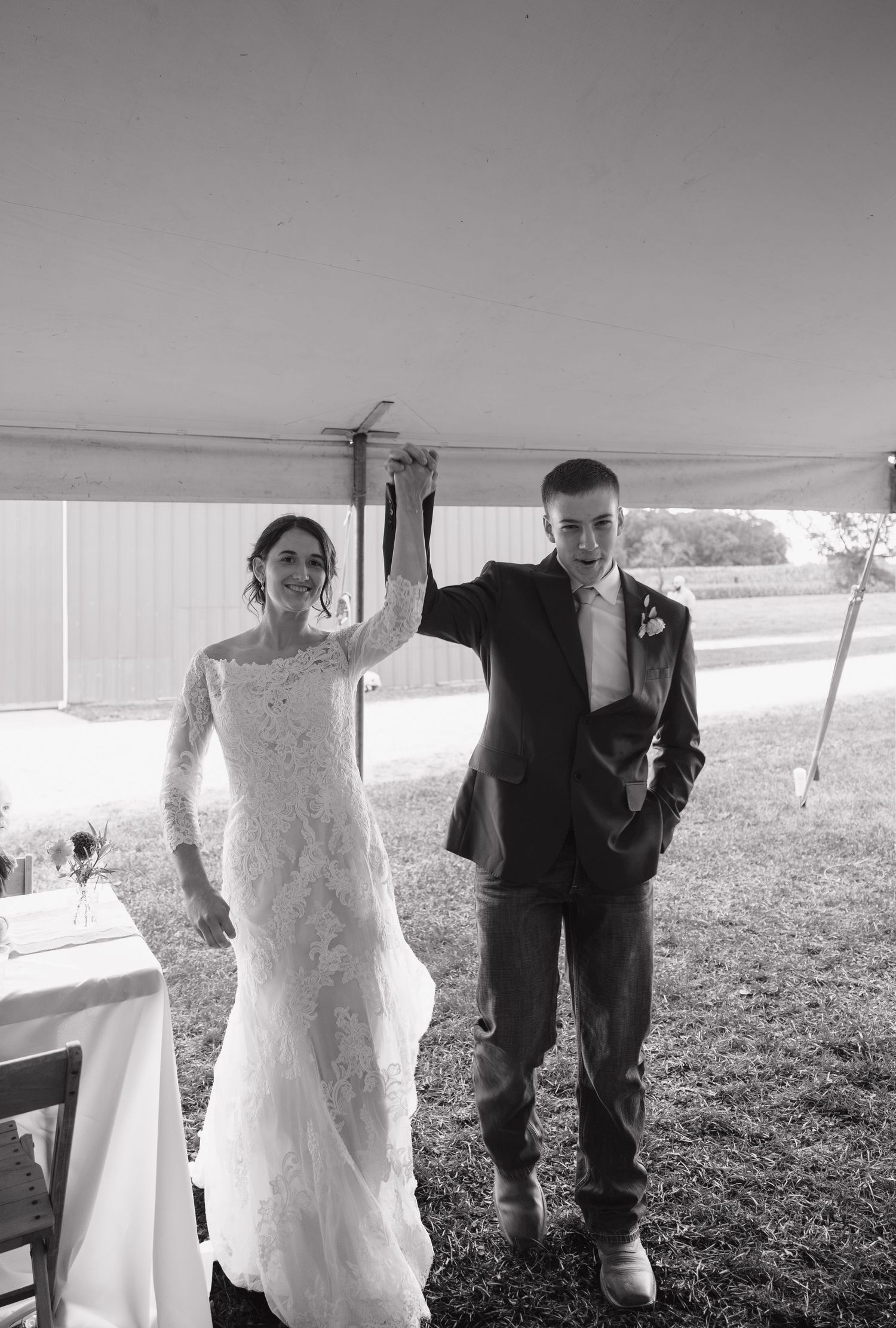 Bride in a lace wedding gown and groom in a suit raise joined hands as they enter a tented outdoor wedding reception in black and white.